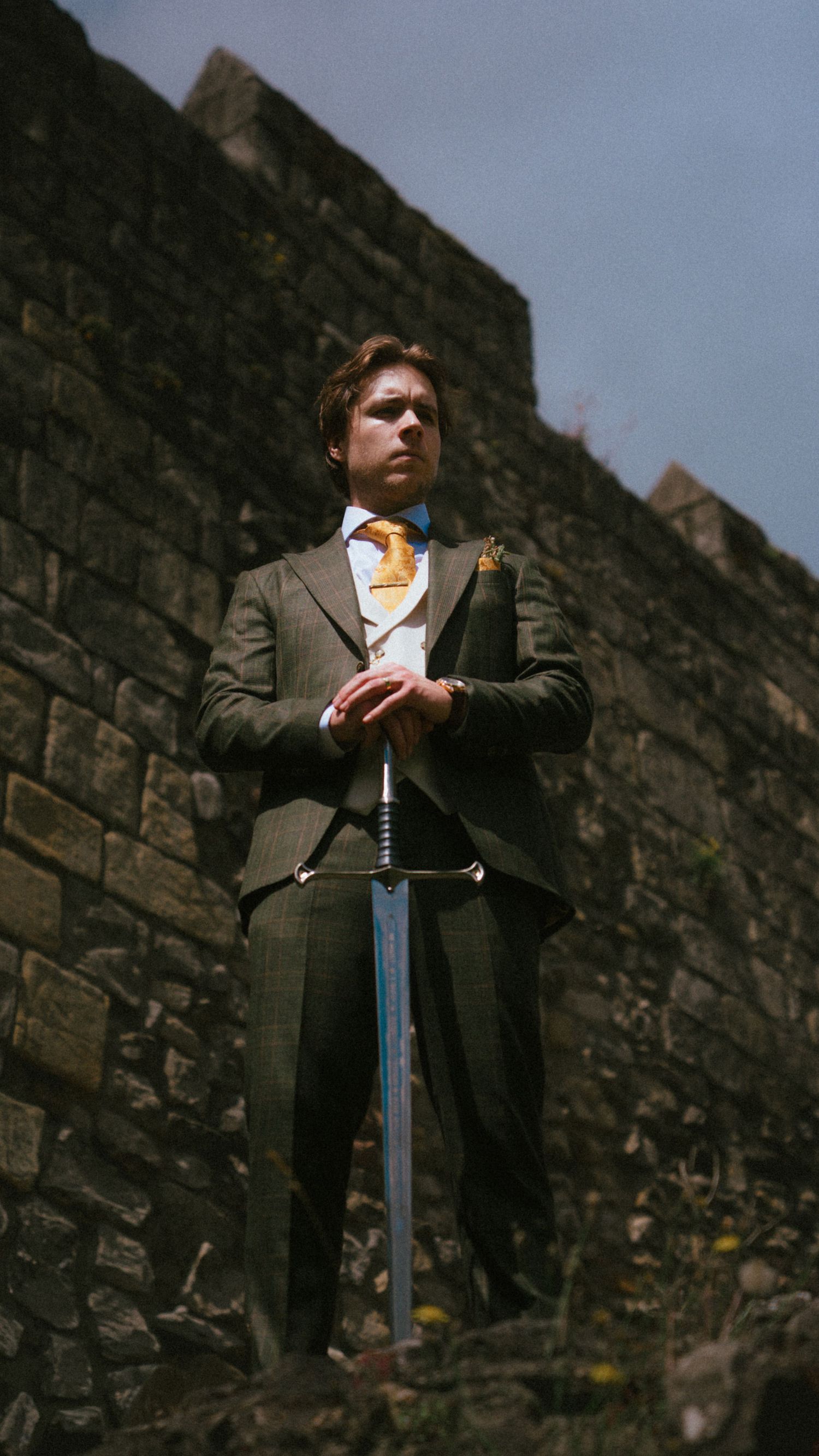 Groom in dark suit holding sword against ancient stone castle wall.