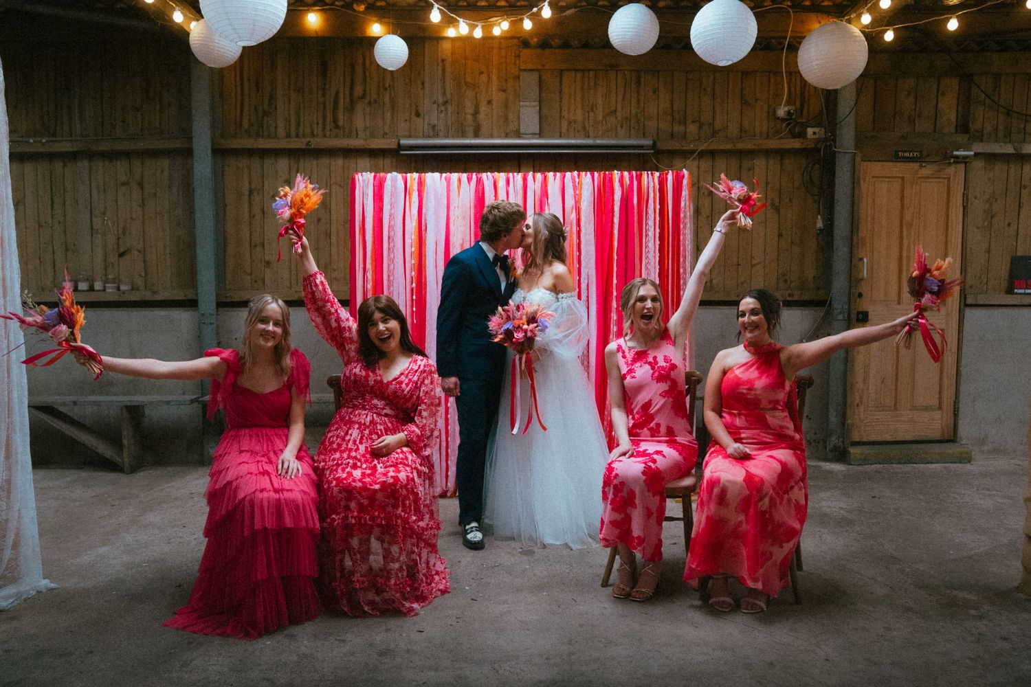 Bride and groom kiss flanked by bridesmaids in pink floral dresses raising bouquets in a rustic barn wedding venue.