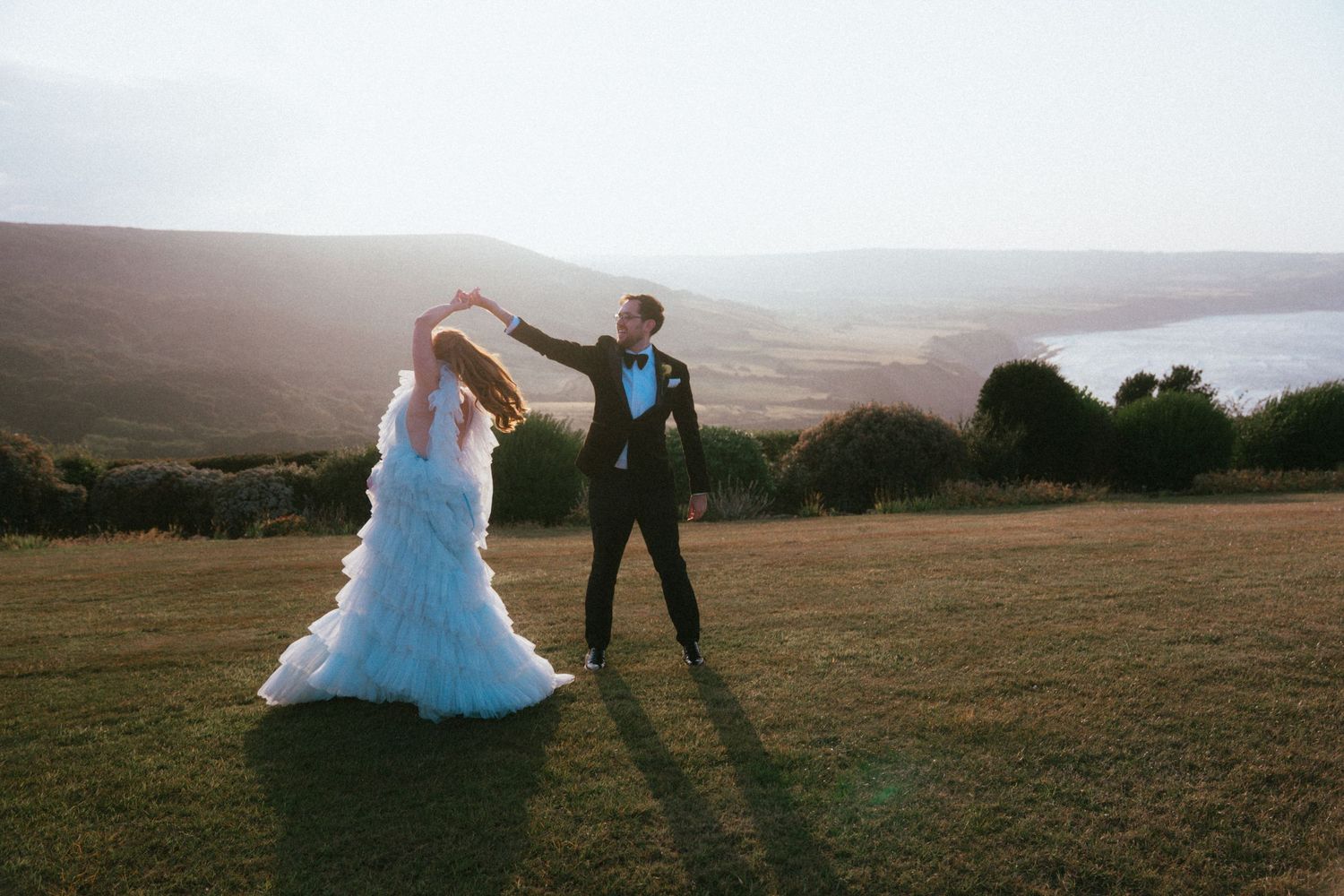 Bride in a flowing white ruffled tulle gown dances with a groom in a dark suit on a grassy hilltop overlooking rolling hills and the sea.