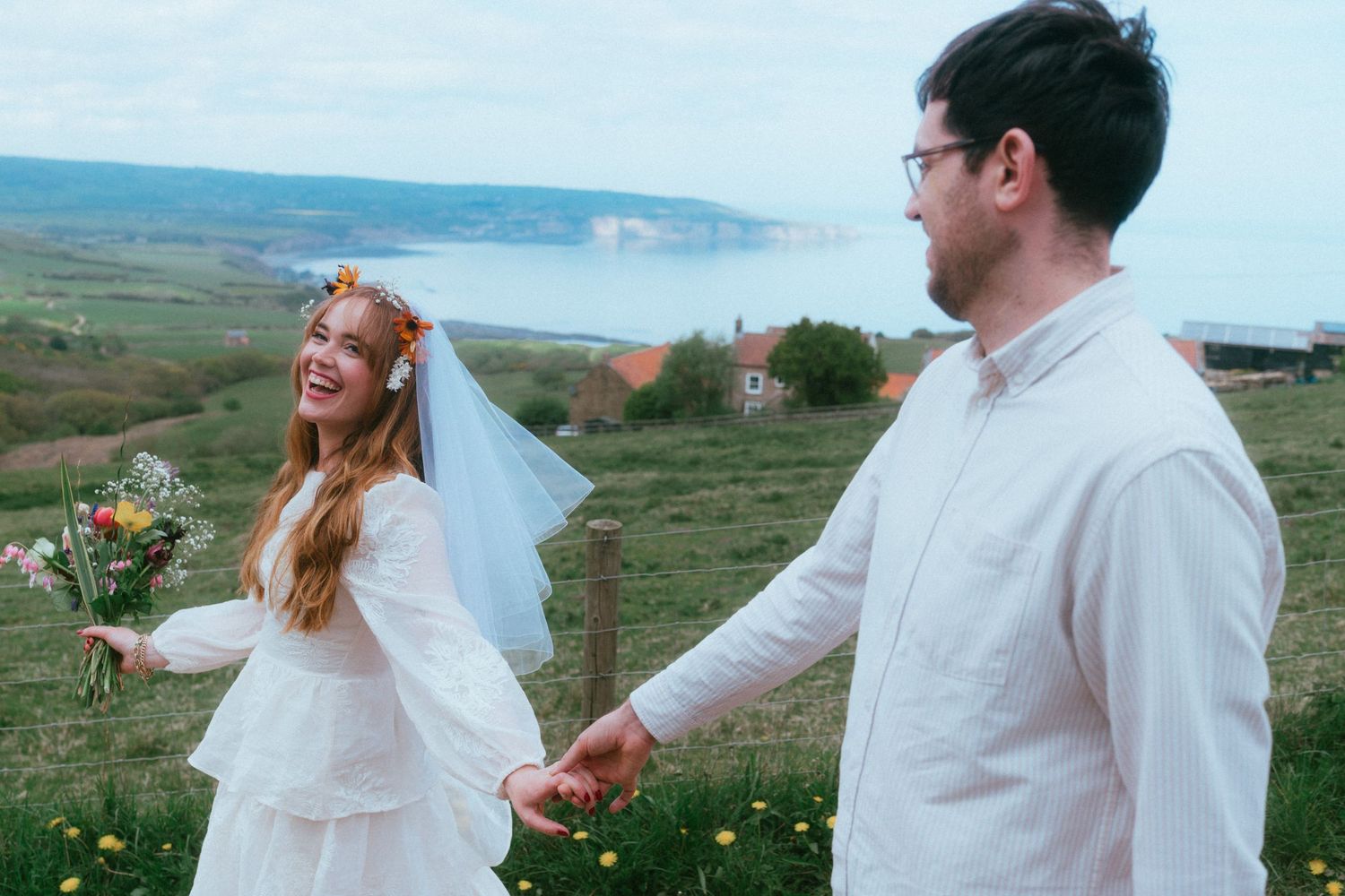 Bride in white dress and floral crown holds bouquet while holding hands with groom outdoors near coastal scenery.