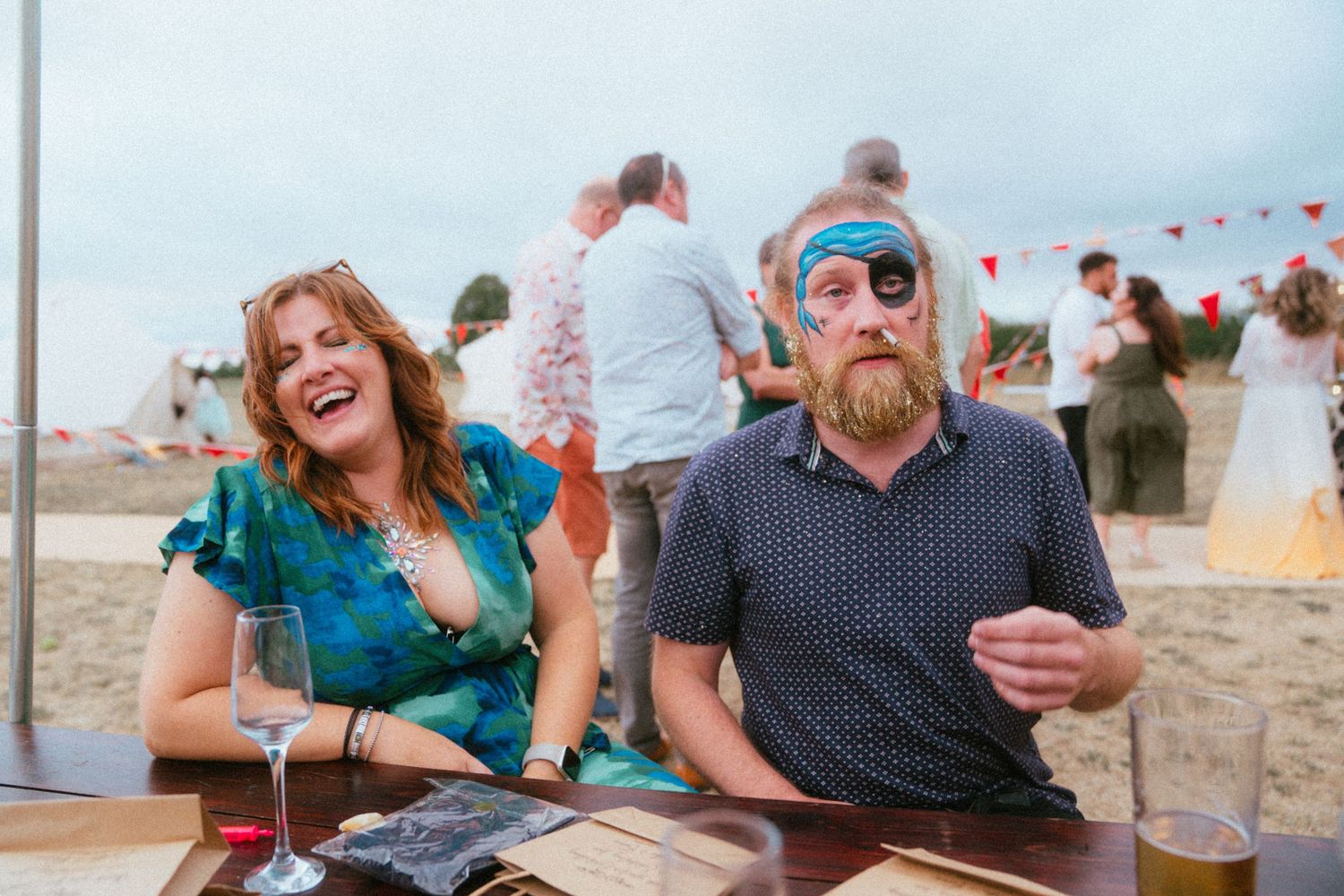 Laughing woman in teal dress sits beside bearded man wearing pirate eye patch at outdoor beach wedding reception.