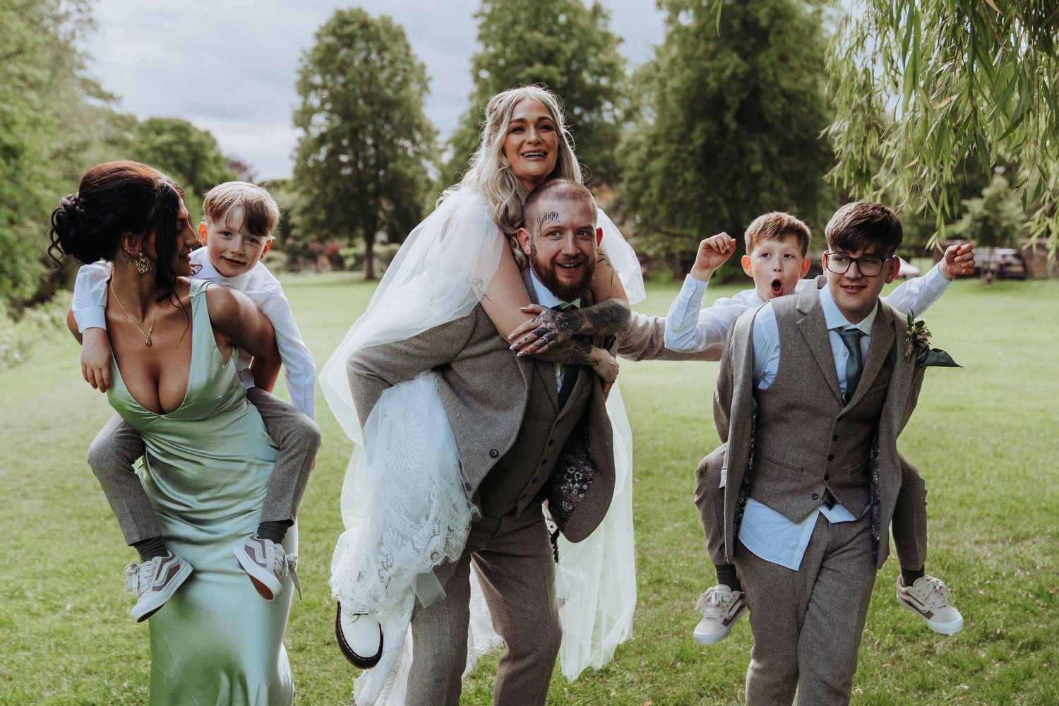 Joyful wedding party photo with bride being carried piggyback by groom while others carry children outdoors.