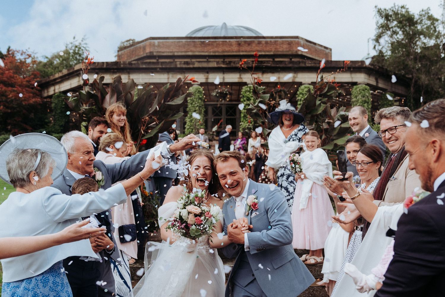 Newlyweds celebrate with confetti shower surrounded by cheering wedding guests outside elegant venue.