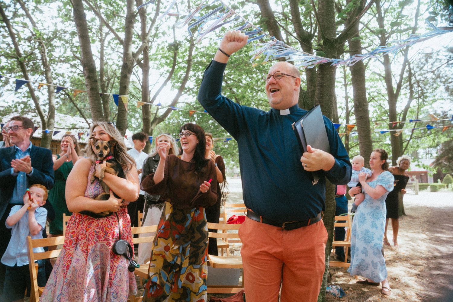 Joyful wedding officiant raises hand in celebration while guests cheer and clap in an outdoor woodland ceremony.