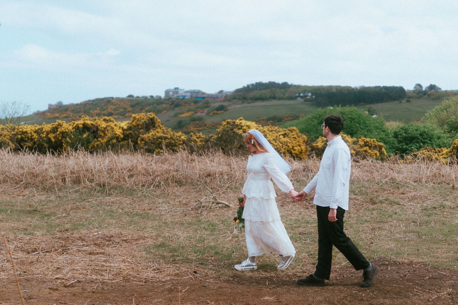 Bride in a white gown and veil walks hand in hand with a groom in a white shirt along a dirt path past golden shrubs and green hills.