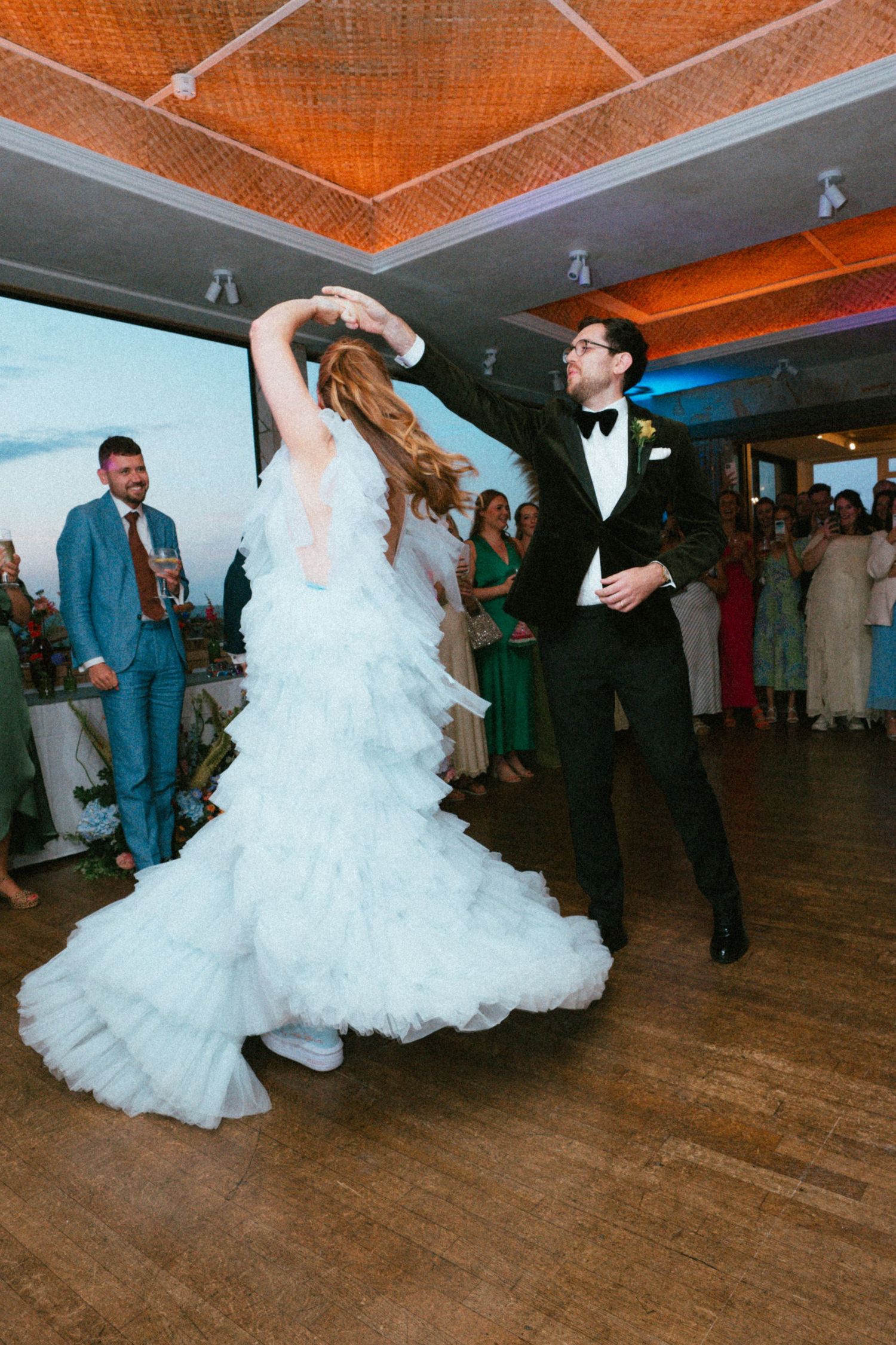 Bride in a flowing light blue tiered ruffle gown twirls with her groom in a black tuxedo during a first dance as wedding guests watch.