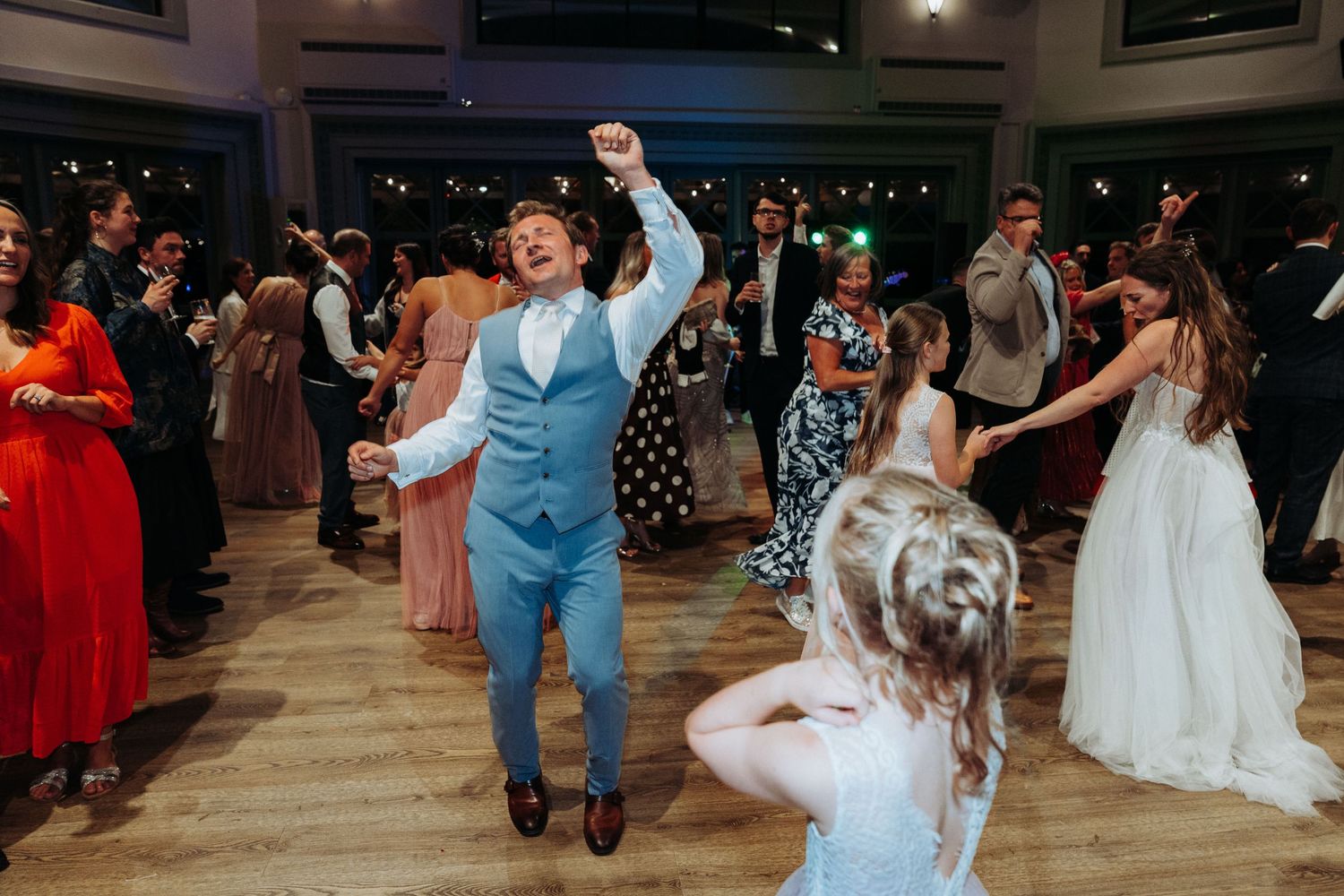 Groom in light blue suit dancing energetically with guests at a lively wedding reception.