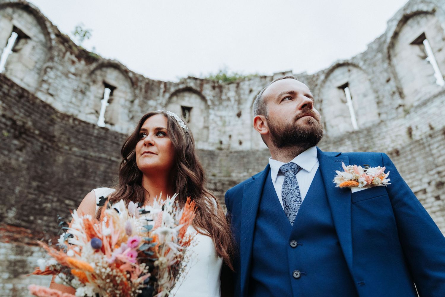 Bride with colorful dried flower bouquet and groom in navy suit pose inside ancient stone ruins.