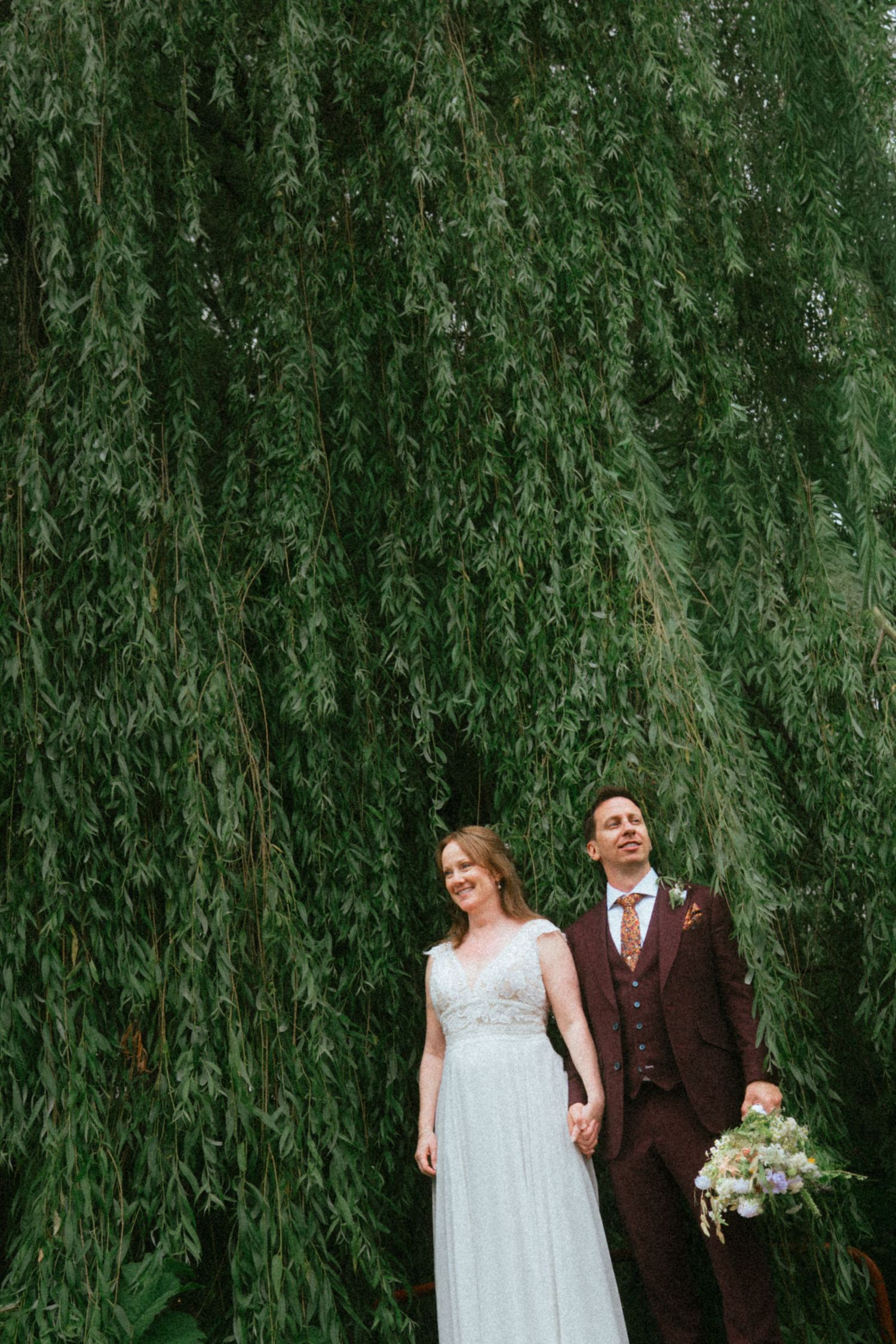 Bride and groom hold hands standing before a lush weeping willow tree on their wedding day.