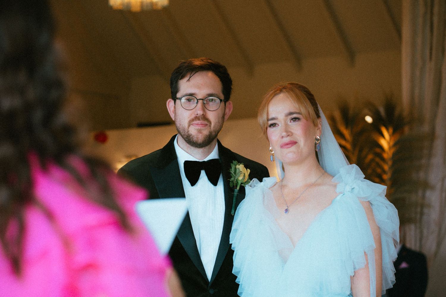 Bride in a light blue tulle gown and veil stands beside a bearded groom in glasses, black tuxedo and bow tie during a wedding ceremony.