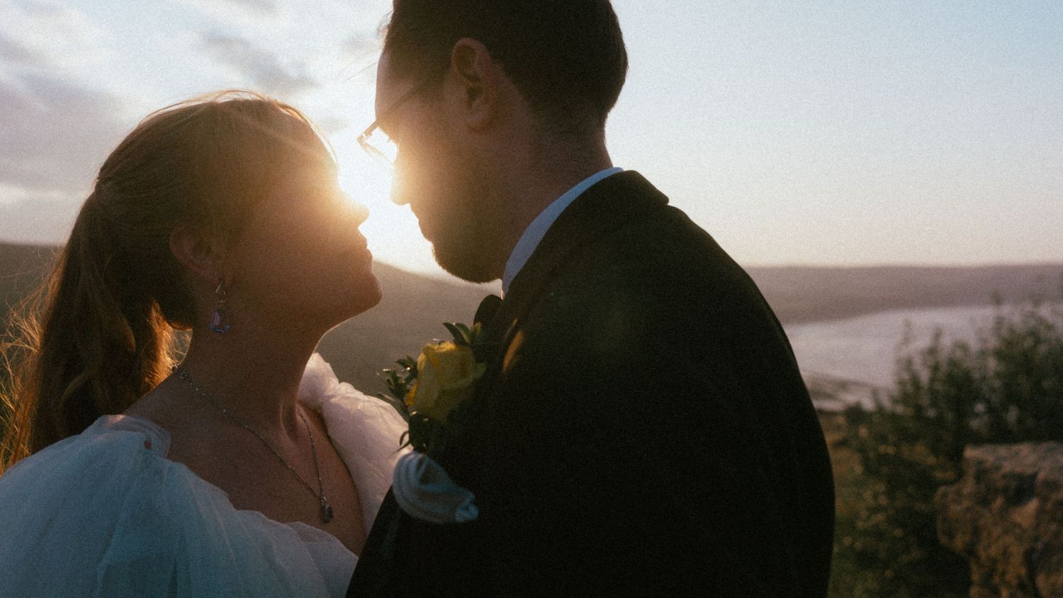 Bride and groom share a tender moment at sunset, faces close together, with golden sunlight glowing behind them and a coastal view beyond.