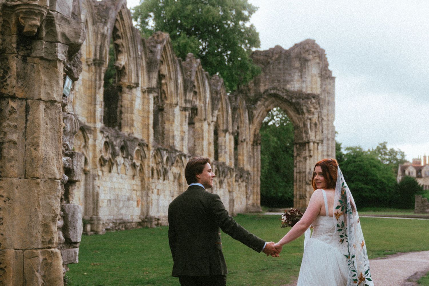 Bride and groom hold hands at ancient stone abbey ruins during romantic outdoor wedding photography session.