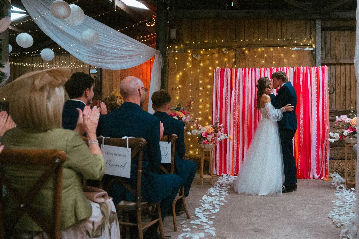Bride and groom share first kiss at rustic barn wedding ceremony with pink ribbon backdrop and fairy lights.