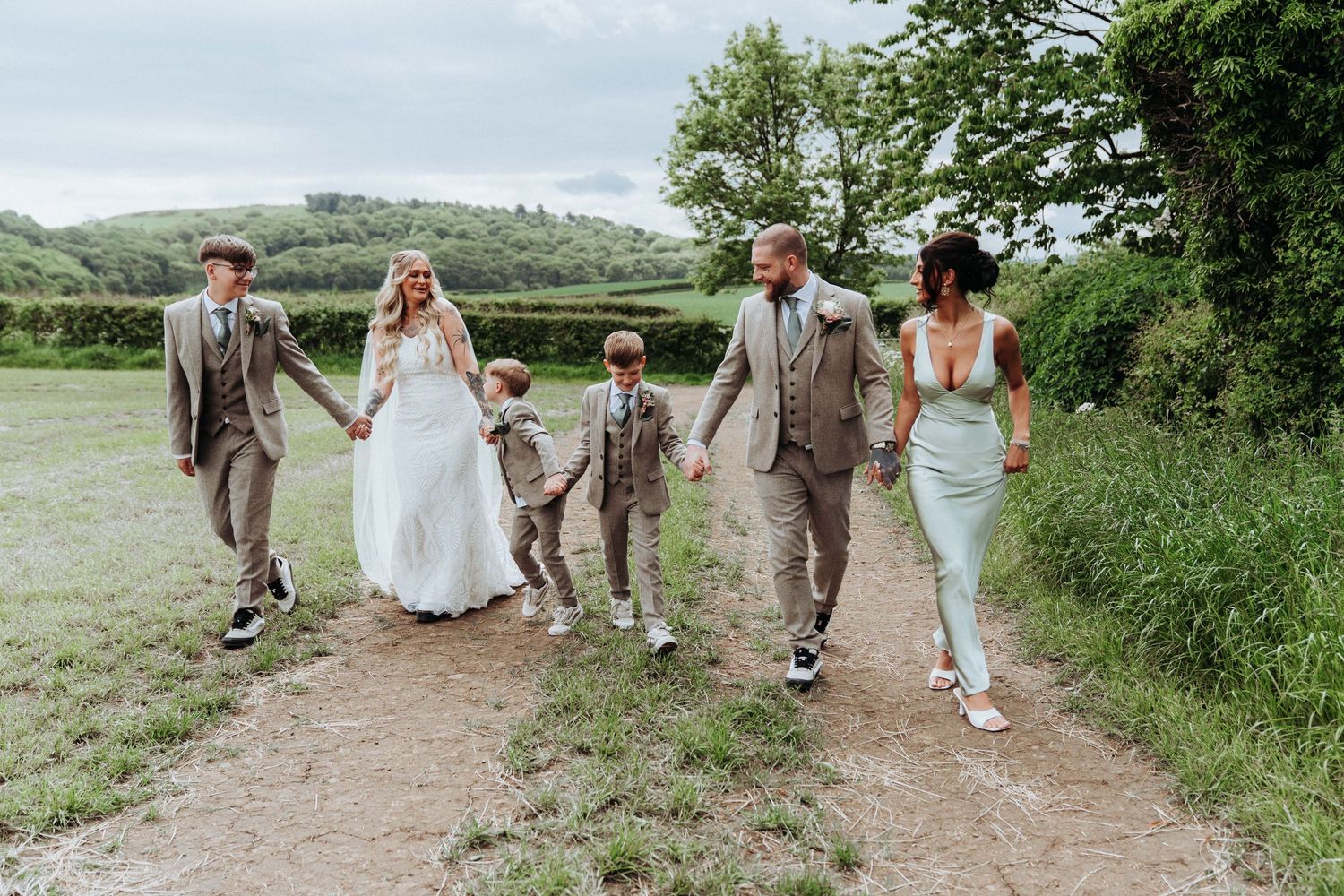 Wedding party walking along countryside path with rolling green hills, bride in white gown with children.
