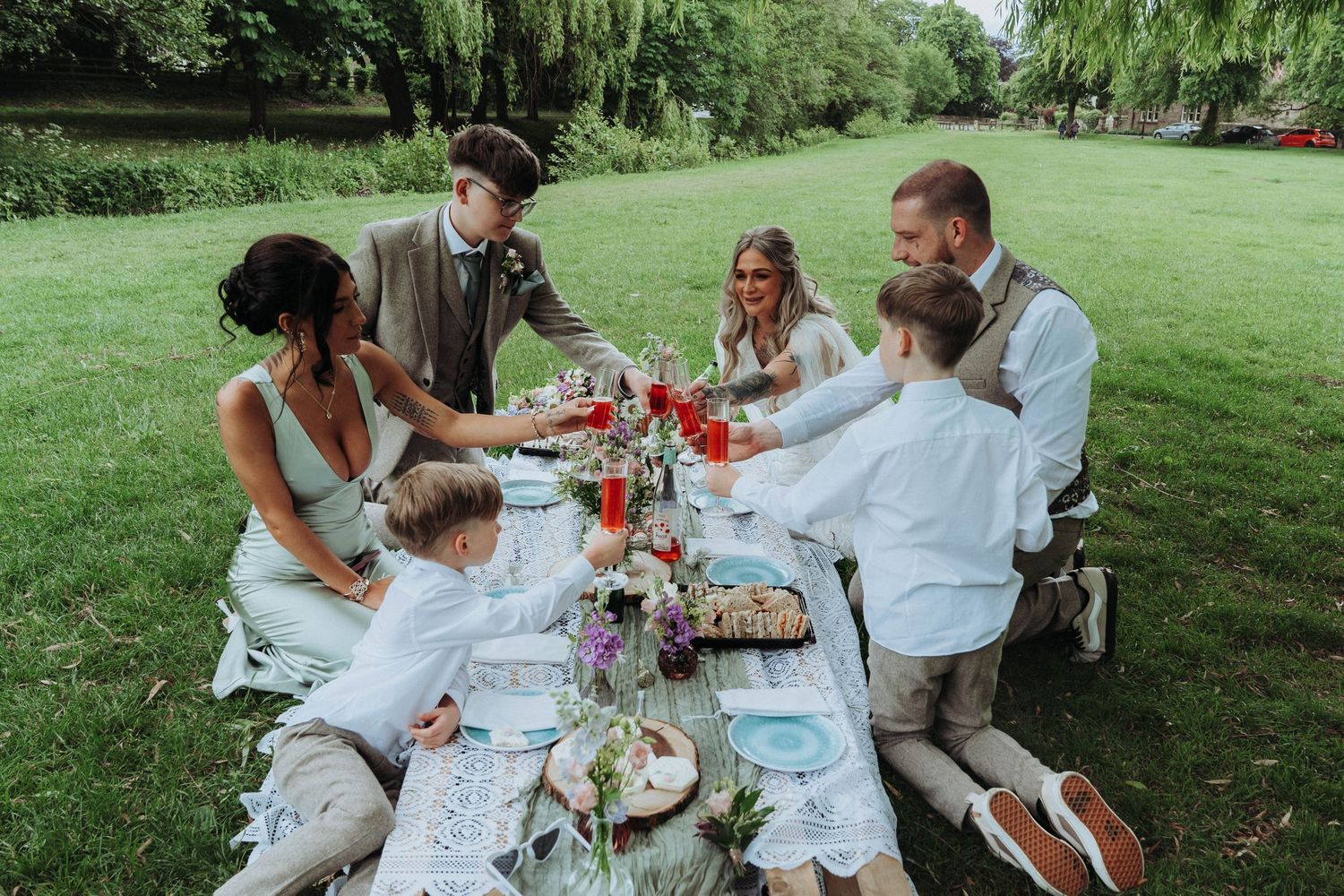 Wedding party enjoying an intimate outdoor picnic reception at a decorated table surrounded by greenery.