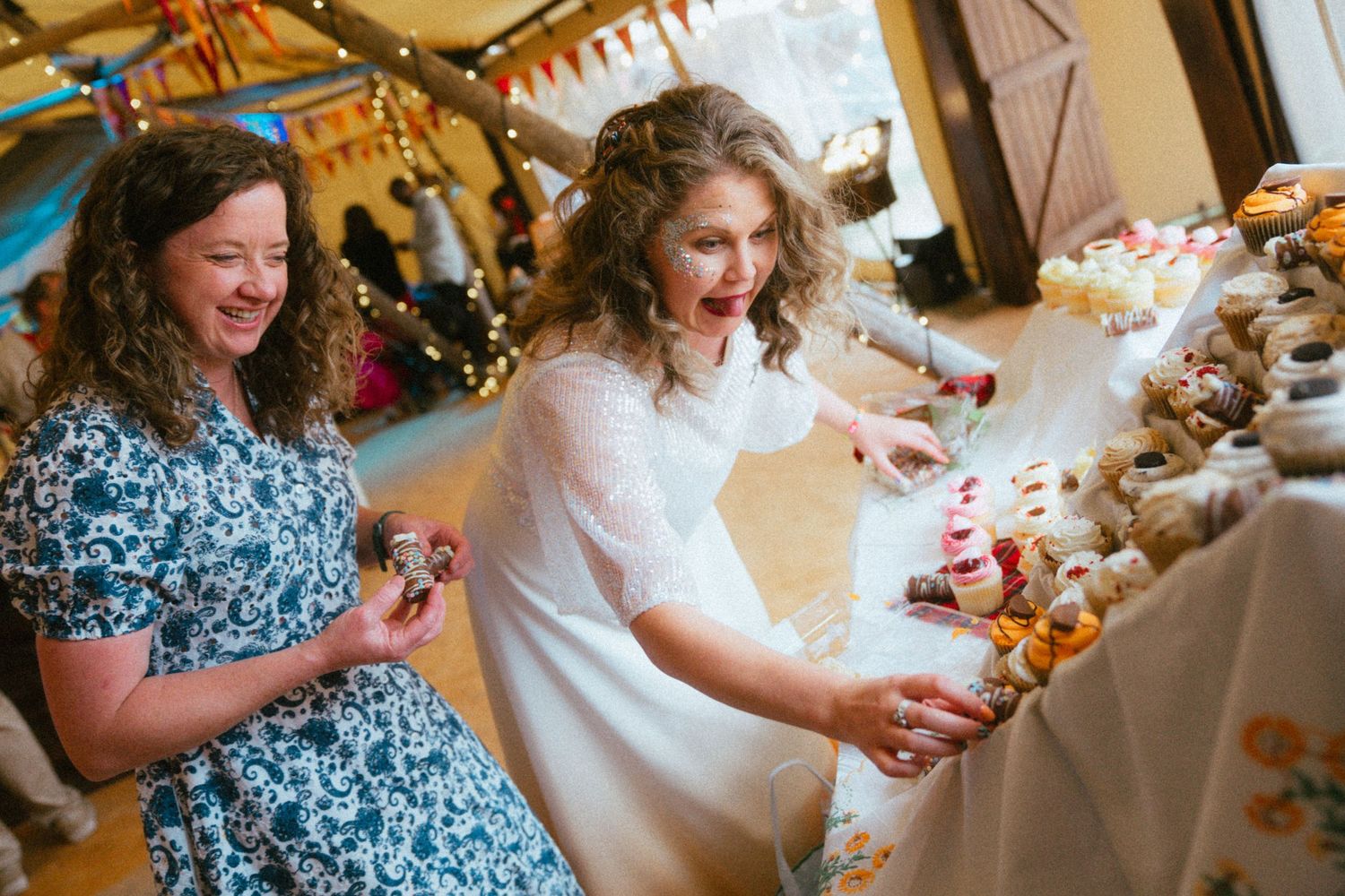 Bride in white dress reaches for cupcakes at a colorful wedding dessert table while a guest in floral dress smiles.