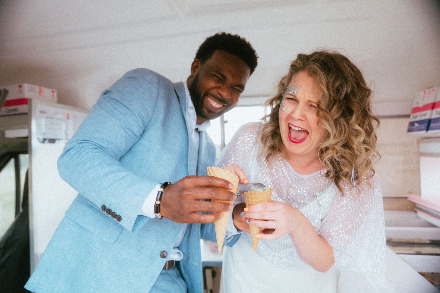 Happy interracial bride and groom share ice cream cones laughing together on their wedding day.