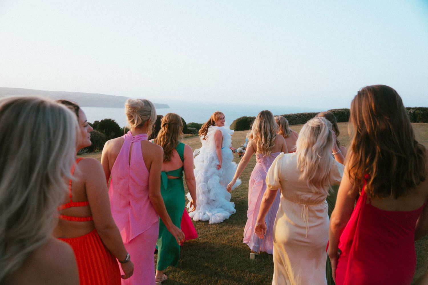 Bridesmaids in colorful pink, orange, green and red dresses walk across a grassy hilltop toward the sea with a bride in a white gown.