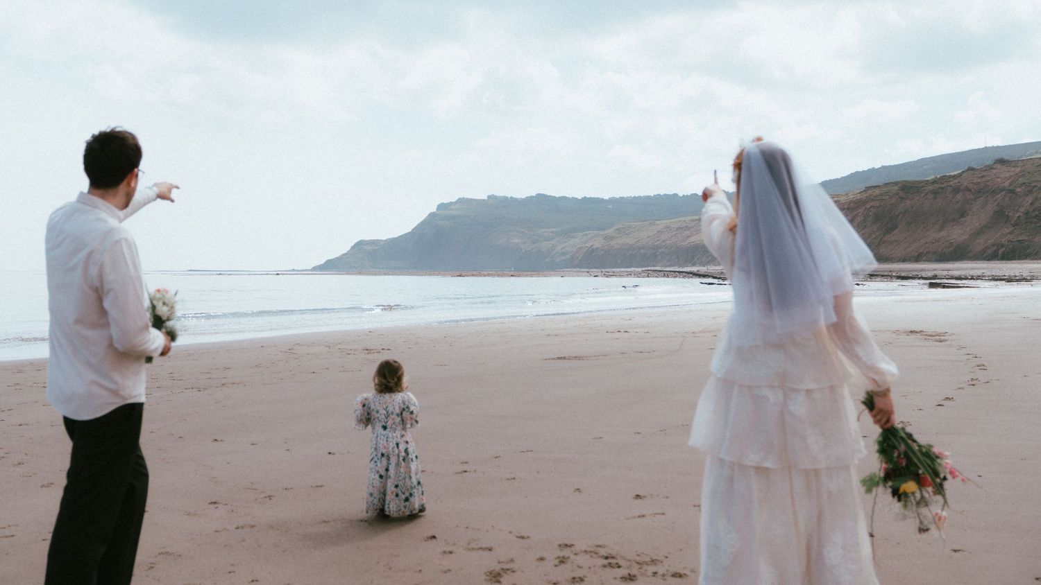 Bride in a white veil and gown and groom in a white shirt stand on a sandy beach with a small child, facing the sea and coastal cliffs.