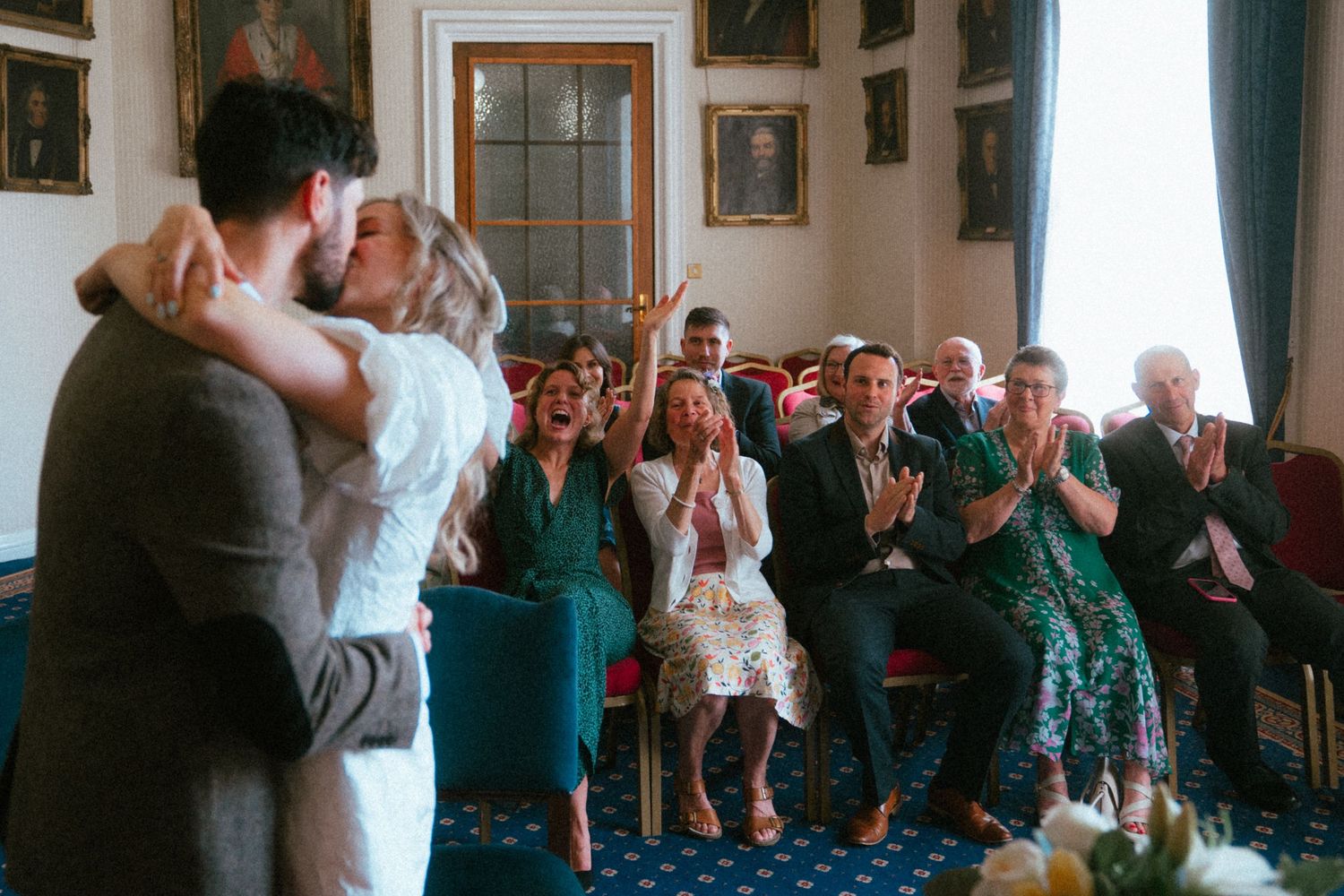 Bride and groom share first kiss at wedding ceremony while guests cheer and applaud in elegant blue-carpeted room.