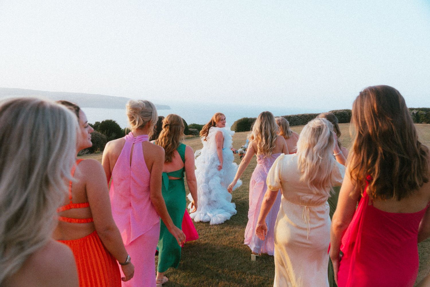 Bridal party in colorful dresses walking across a sunny field toward a bride in a white gown.