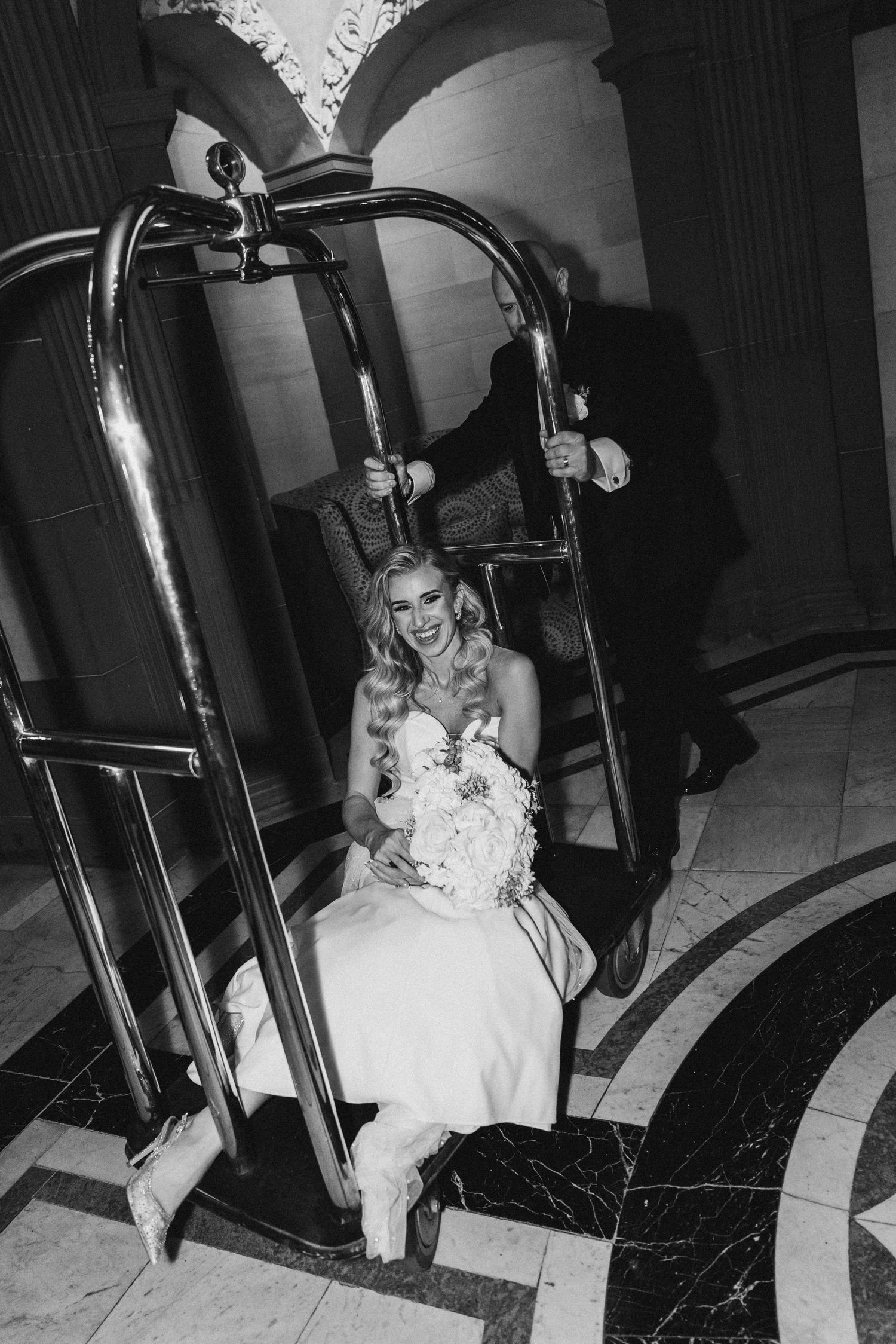 Laughing bride in a wedding gown sits in a hotel luggage cart holding a bouquet.