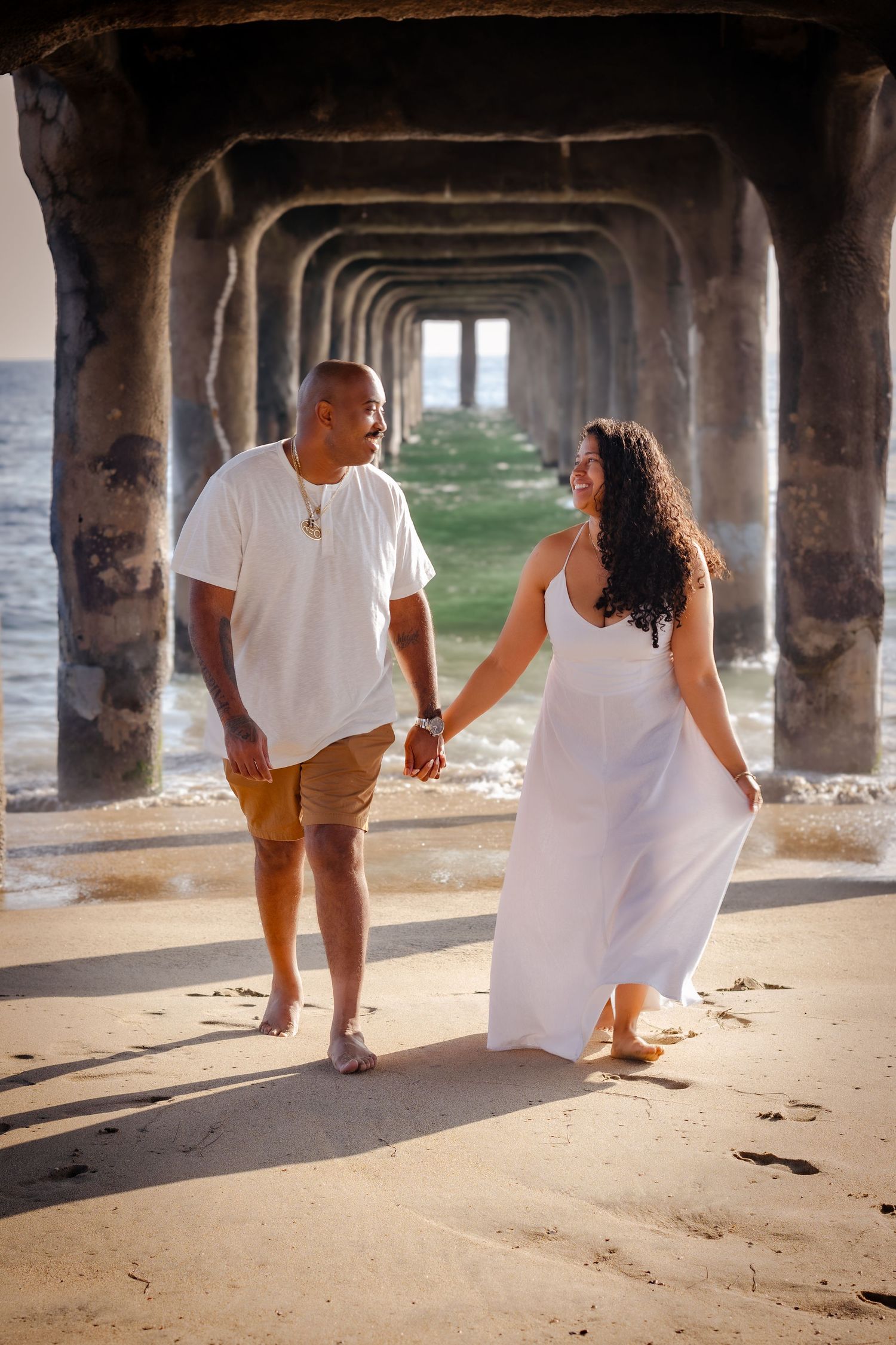 A couple in white outfits walking playfully under a beach pier during sunset, creating a romantic seaside moment.