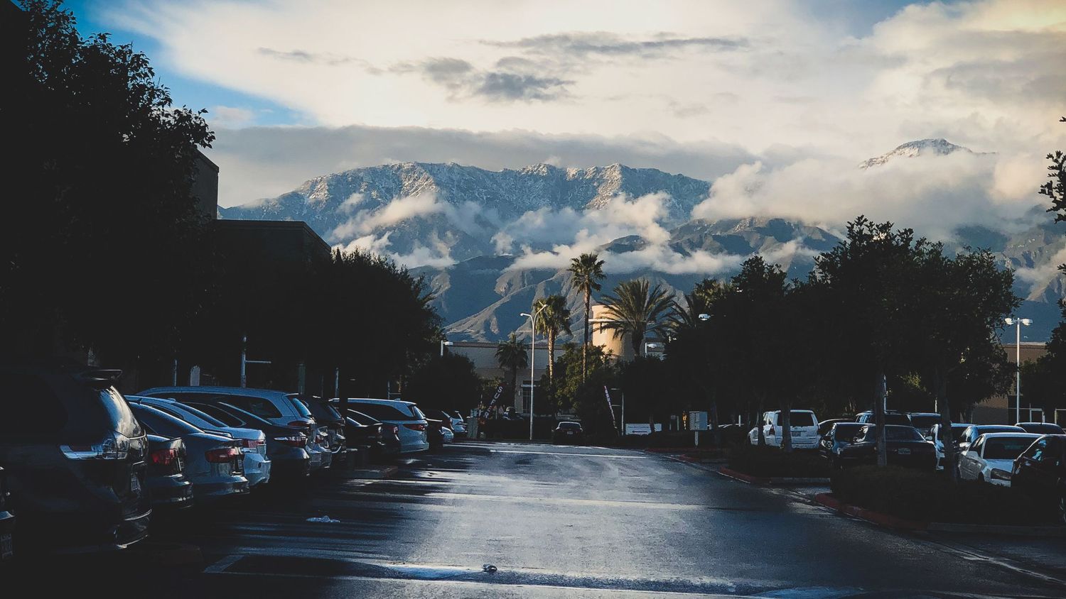 A panoramic view of mountains and clouds at sunset with parked cars and buildings silhouetted in the foreground.