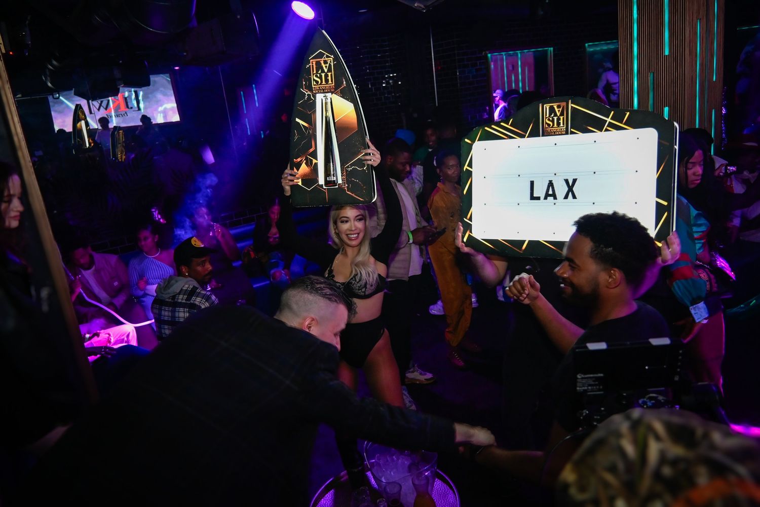 Concert crowd holds up glowing phones during a performance in a dark venue with purple lighting effects.