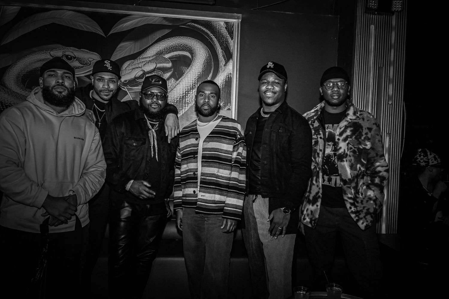 A black and white photograph shows a group of six people standing together in casual attire against a dark background.