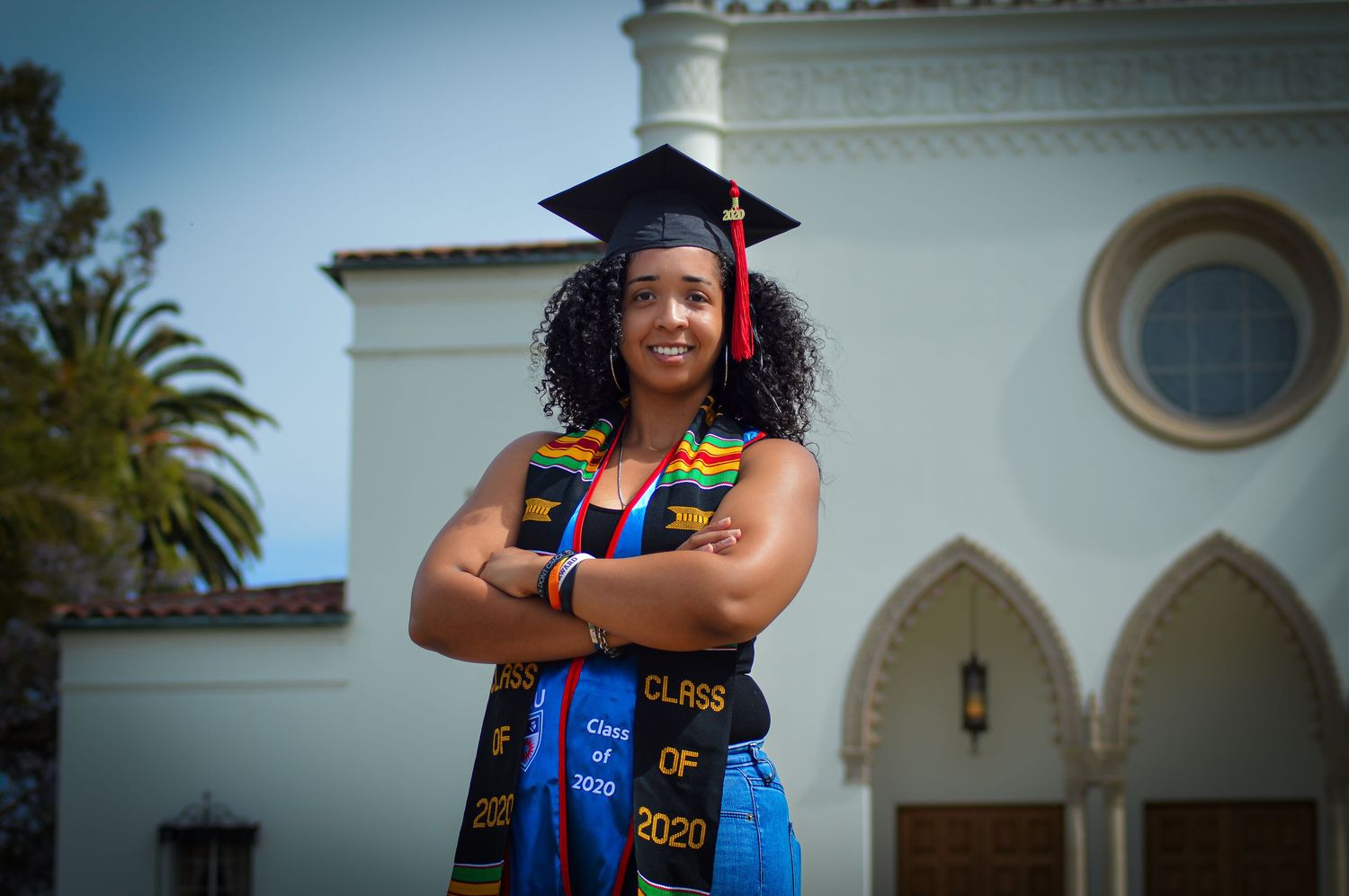 Graduate in blue and gold regalia poses in front of white building with palm trees during commencement ceremony.