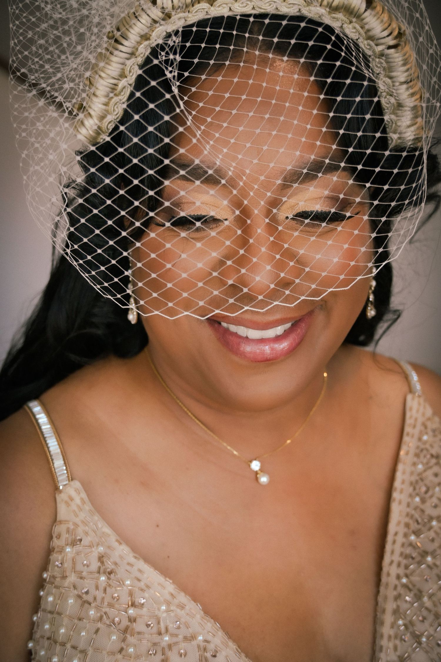 Smiling bride wearing a birdcage veil, pearl earrings, gold necklace, and beaded gown in an elegant bridal portrait.