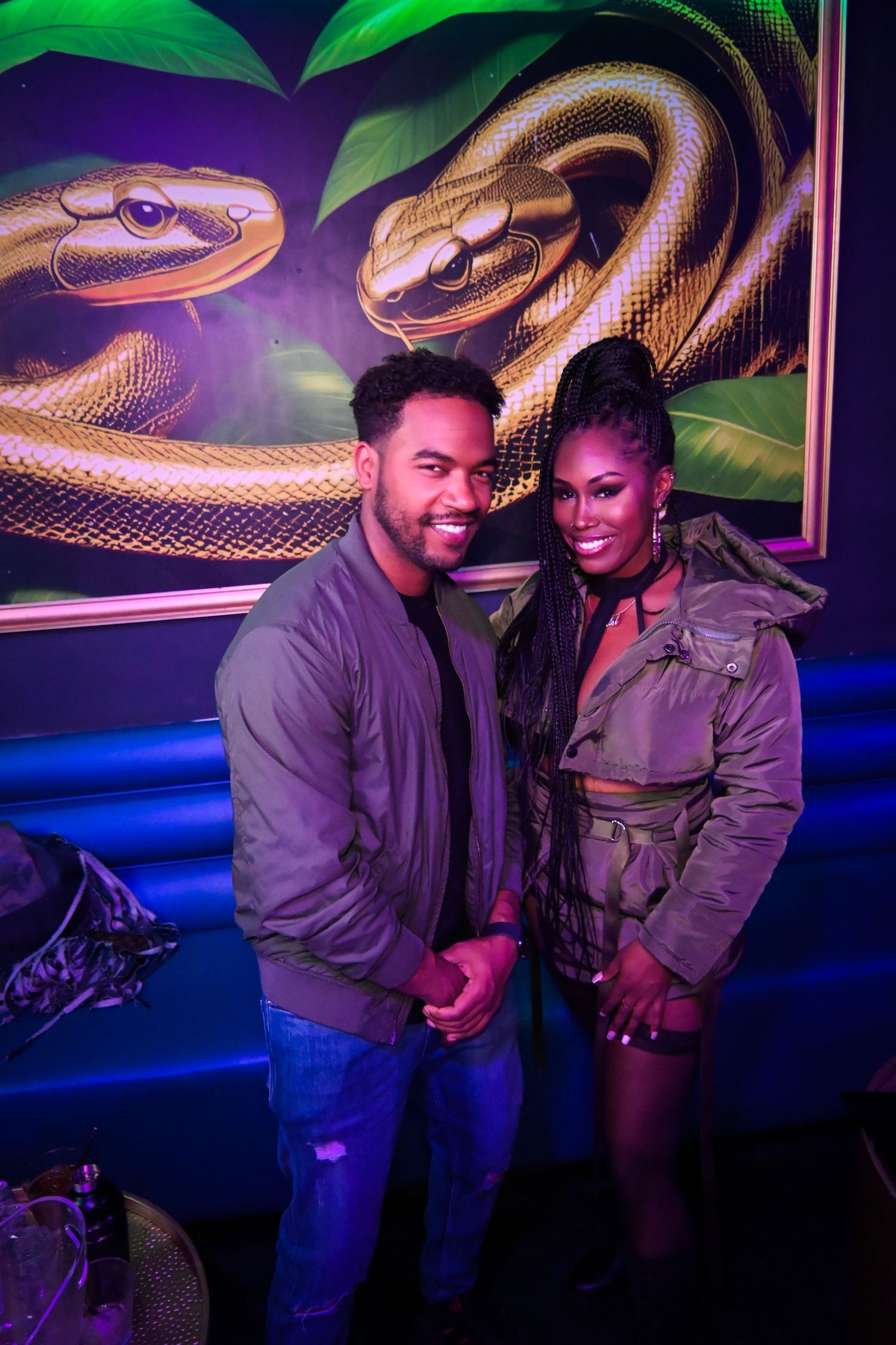 Two people pose together at a nightclub with colorful neon snake artwork glowing on the wall behind them.