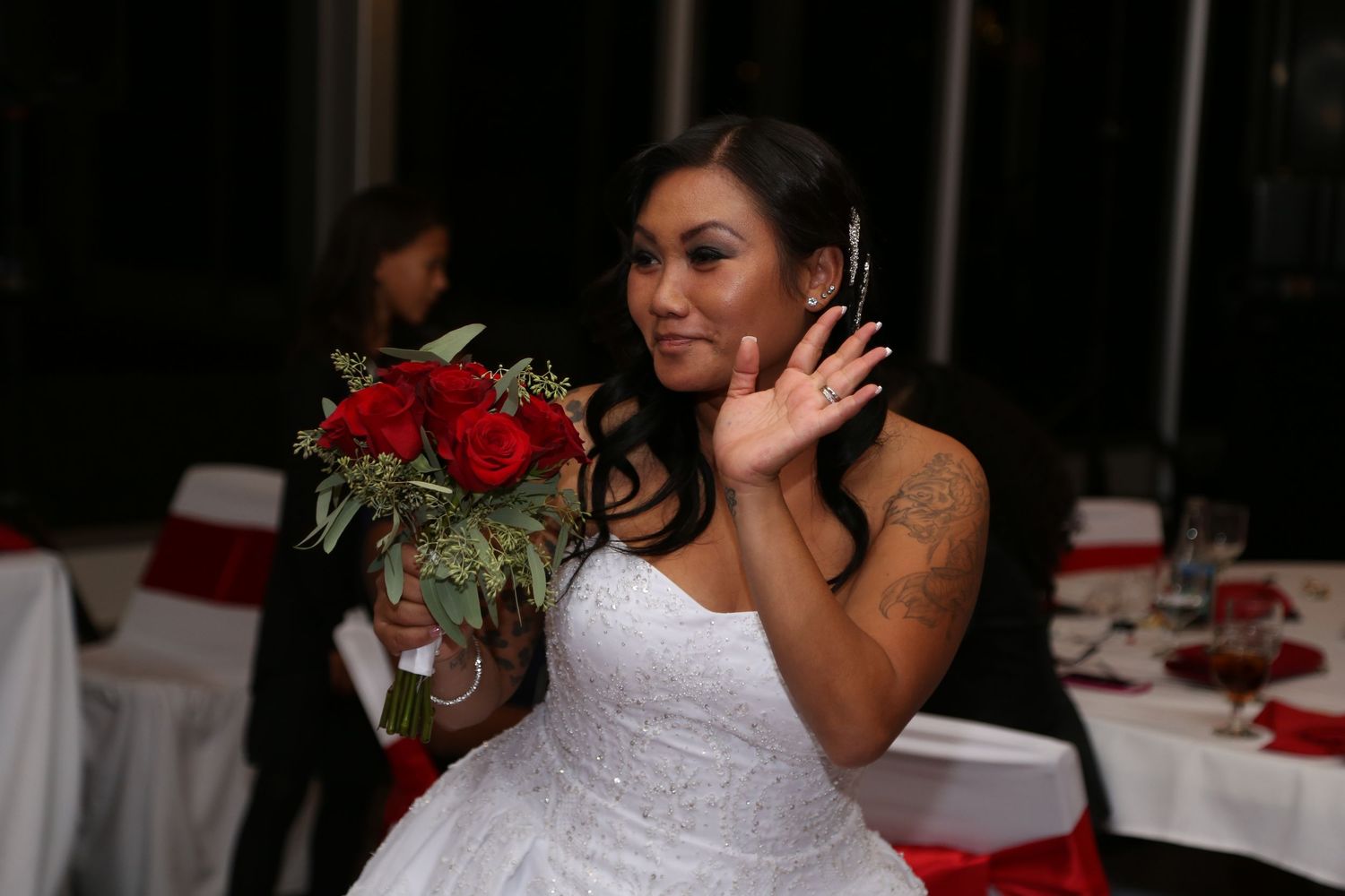 Bride in white strapless gown holding red rose bouquet waves at wedding reception.