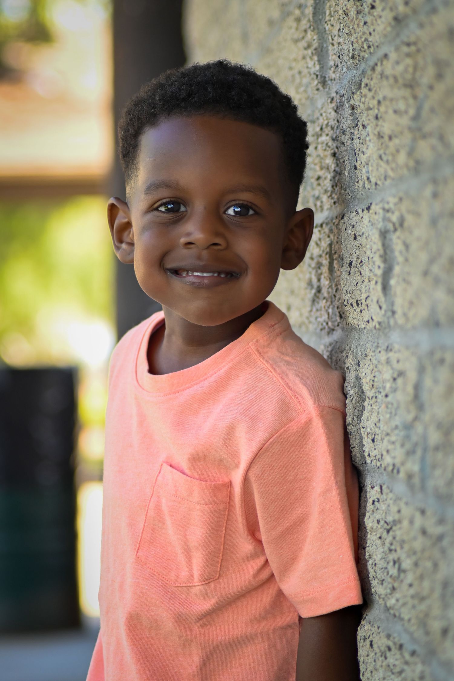 Smiling young Black boy in a coral t-shirt leaning against a stone wall outdoors.