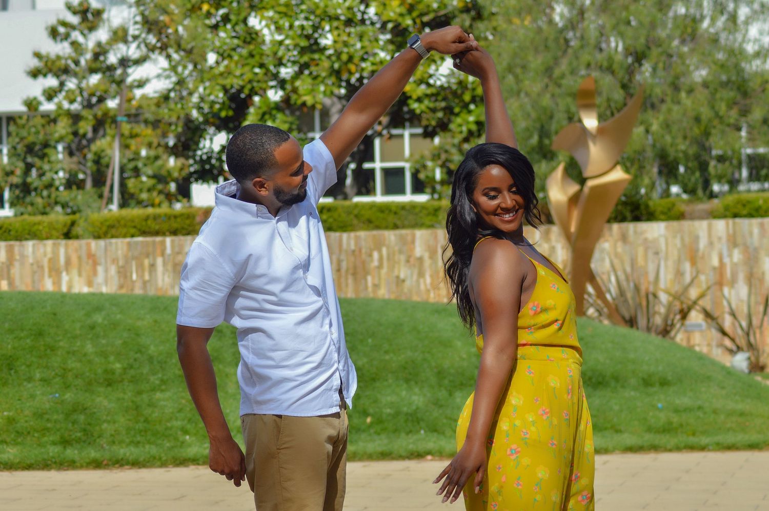 A couple dancing outdoors, man in blue shirt spinning woman in yellow dress, with green lawn and building in background.