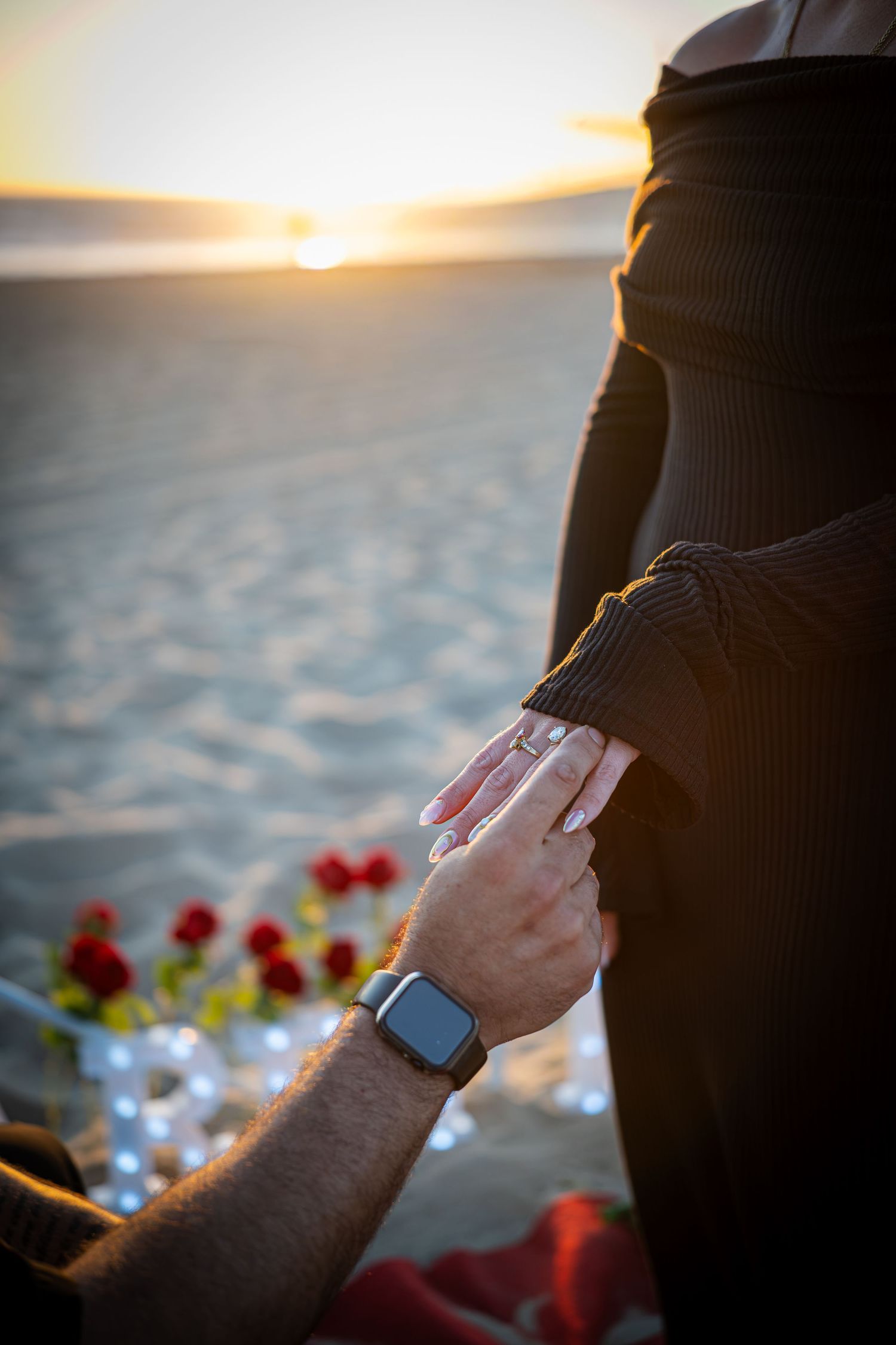 Man placing engagement ring on woman's finger during romantic beach sunset proposal with red roses.