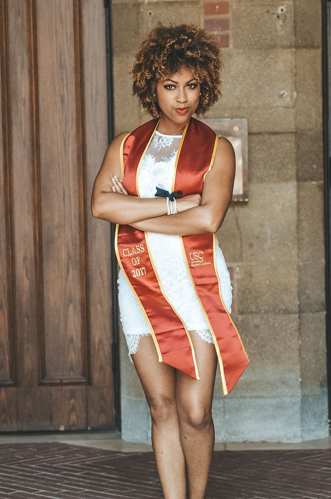 Graduate in white lace dress and red graduation stole poses confidently against stone wall.