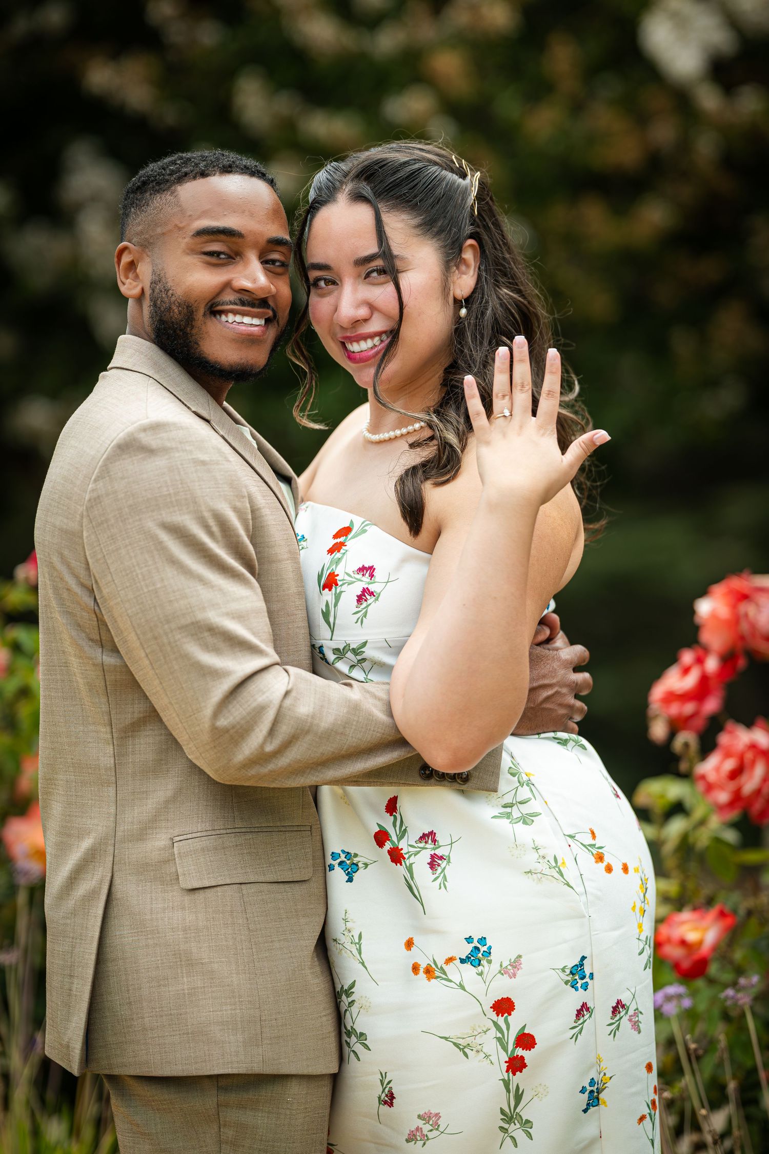 Happy couple poses in garden, woman showing engagement ring, man in tan suit, floral dress, roses in background.