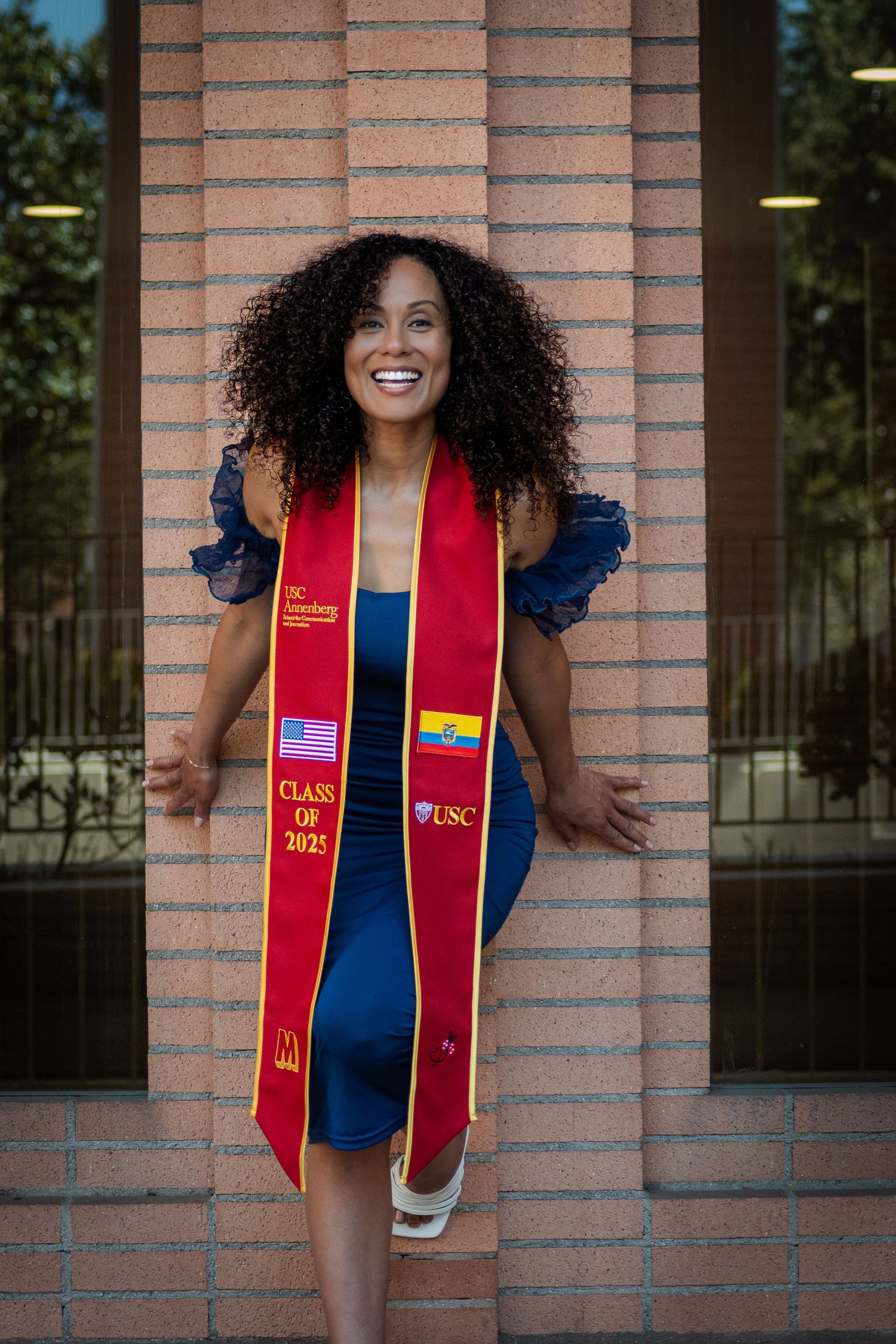 Graduate in blue dress and red graduation stole smiling joyfully against brick wall background.