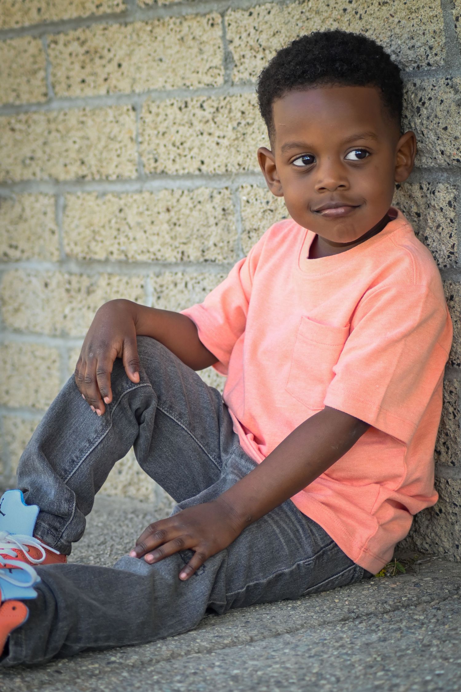 Young Black boy in orange shirt and jeans sits against a brick wall, looking thoughtfully at the camera.