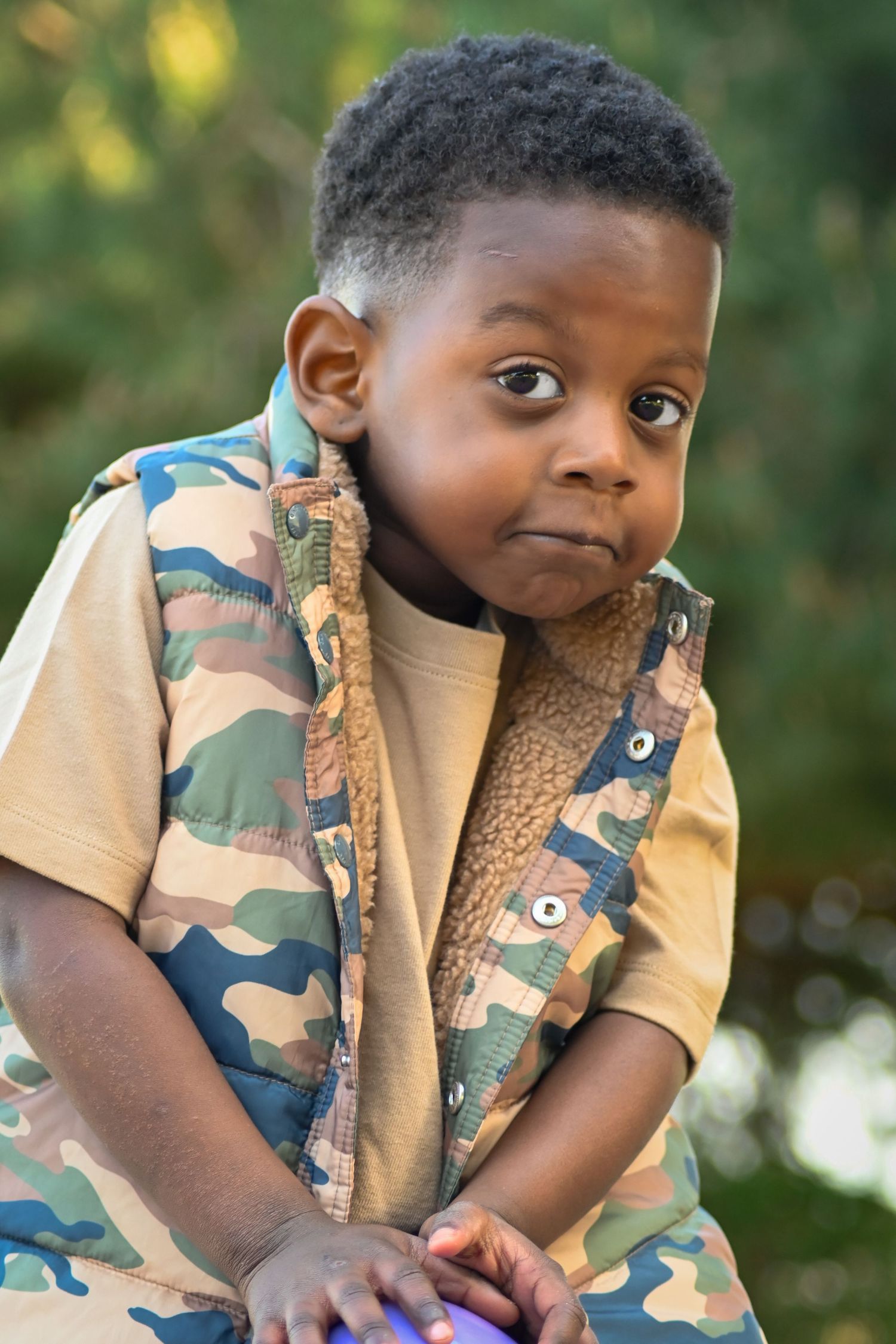 Young Black boy in camouflage vest sitting outdoors, looking at camera with a thoughtful expression.