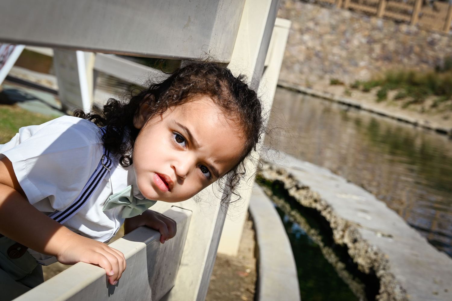 Young girl with curly hair leans over a white bridge railing near a pond, wearing a white sailor-style outfit.