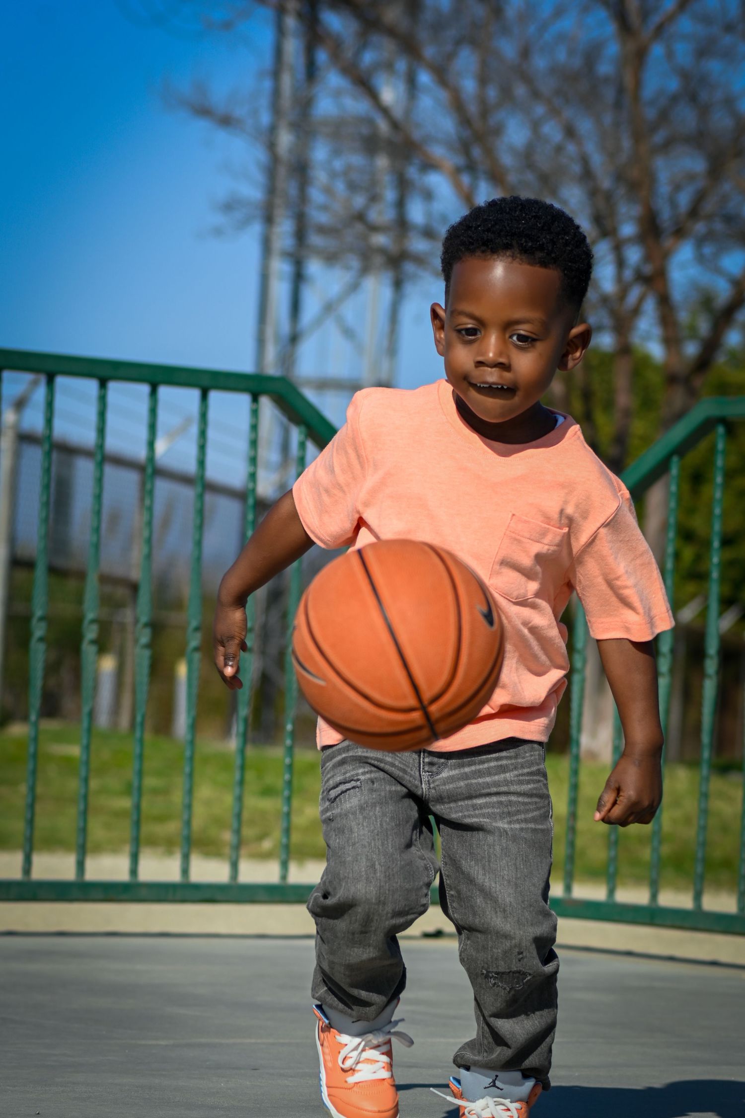 Young Black boy dribbling a basketball on an outdoor court, wearing a peach shirt and jeans on a sunny day.