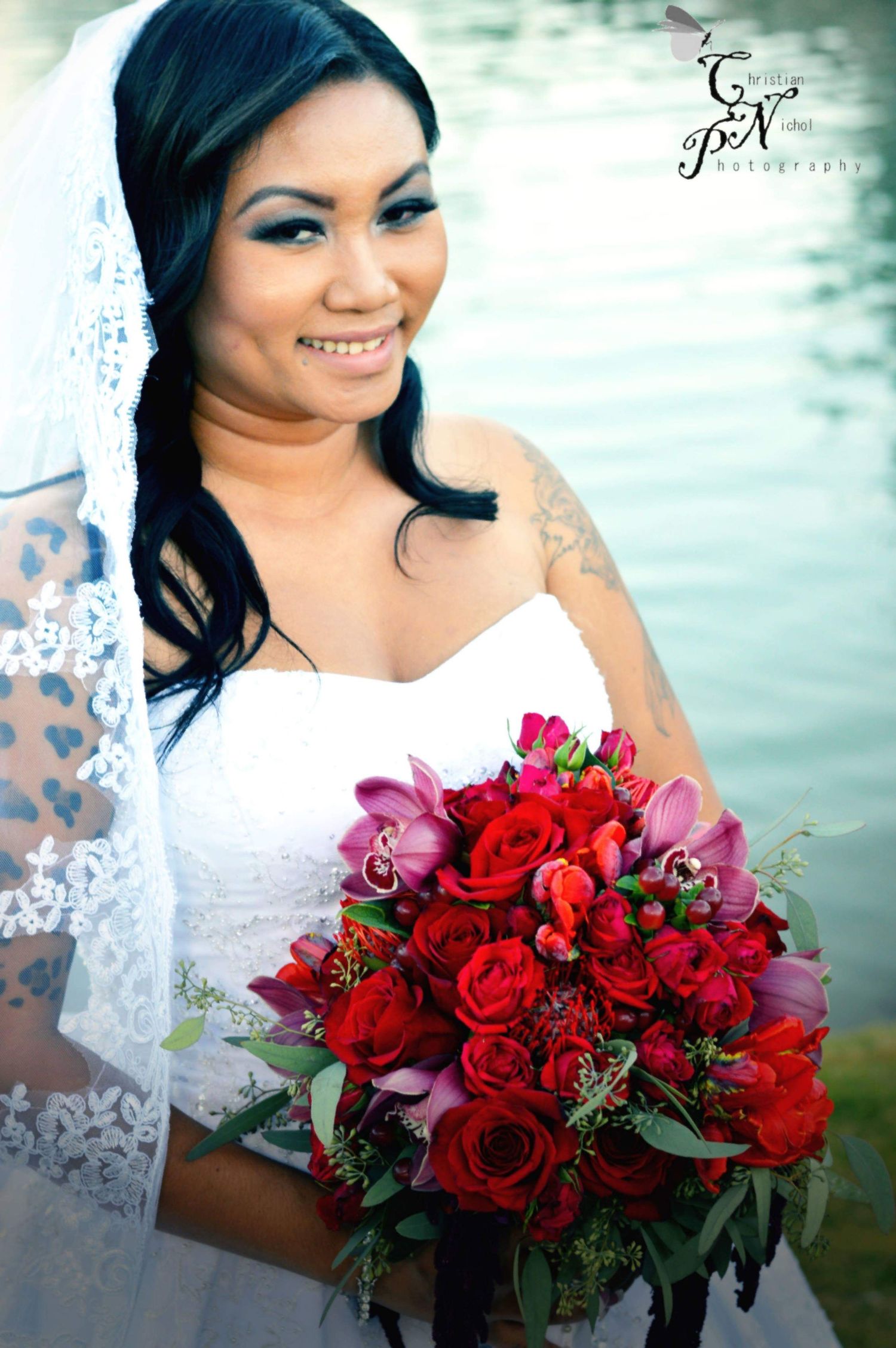 Smiling bride in white lace gown and veil holds vibrant red rose and pink orchid bouquet outdoors near water.