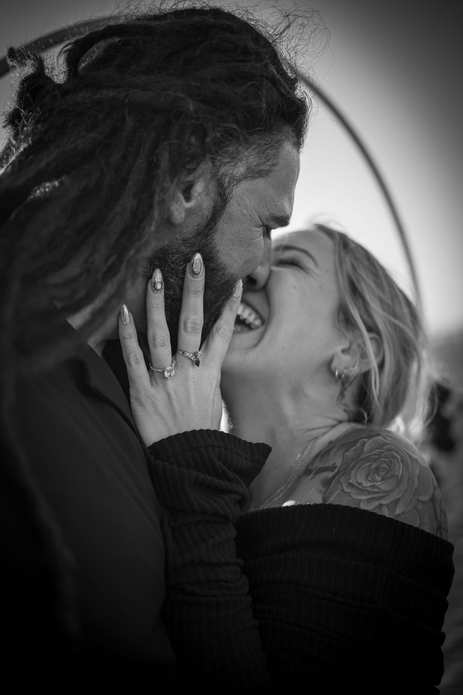 Black and white photo of a laughing couple sharing a romantic moment, woman with tattoo and rings, man with dreadlocks.