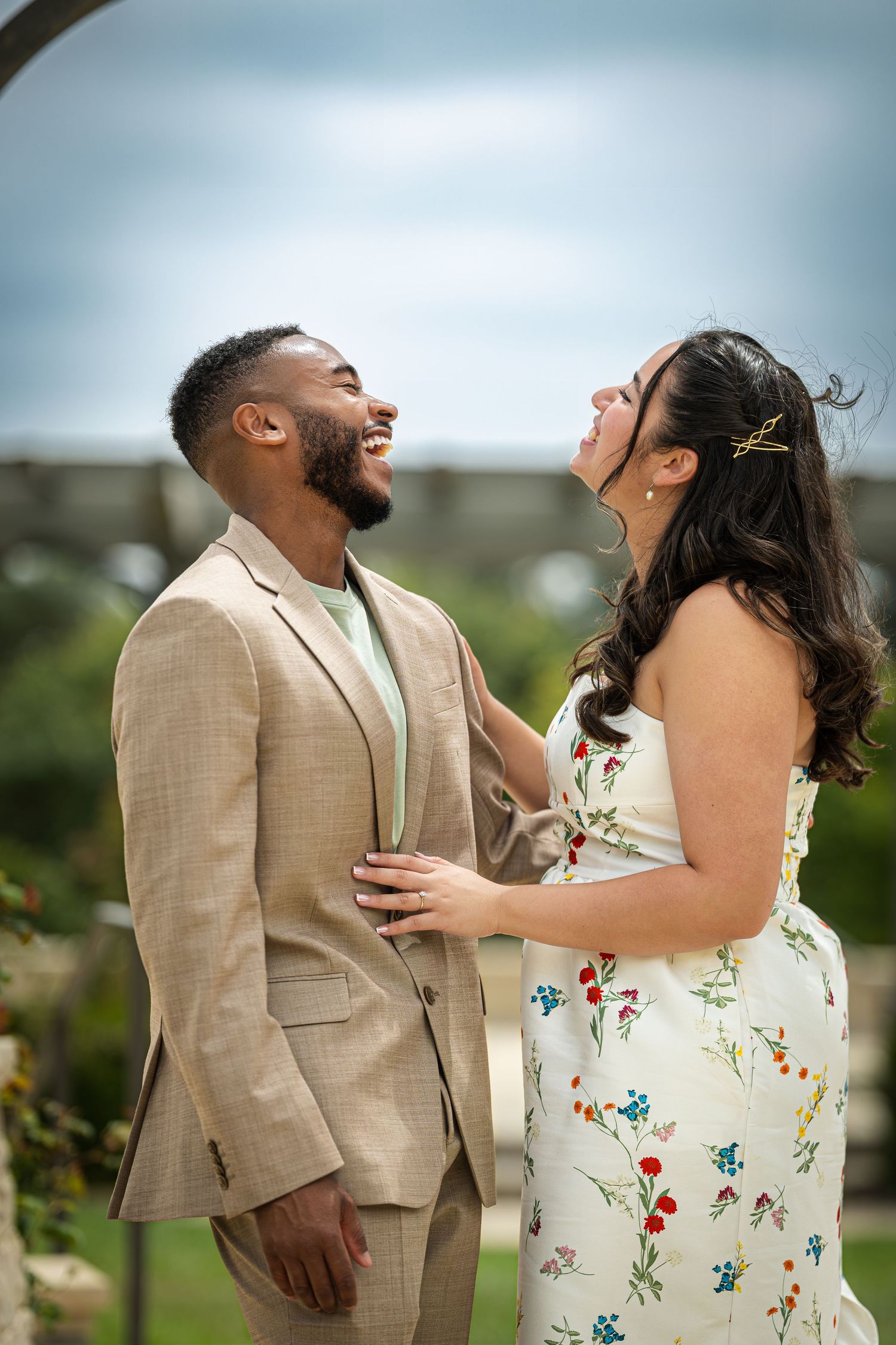 A joyful couple laughs together outdoors, man in tan suit, woman in floral dress, at a garden wedding ceremony.