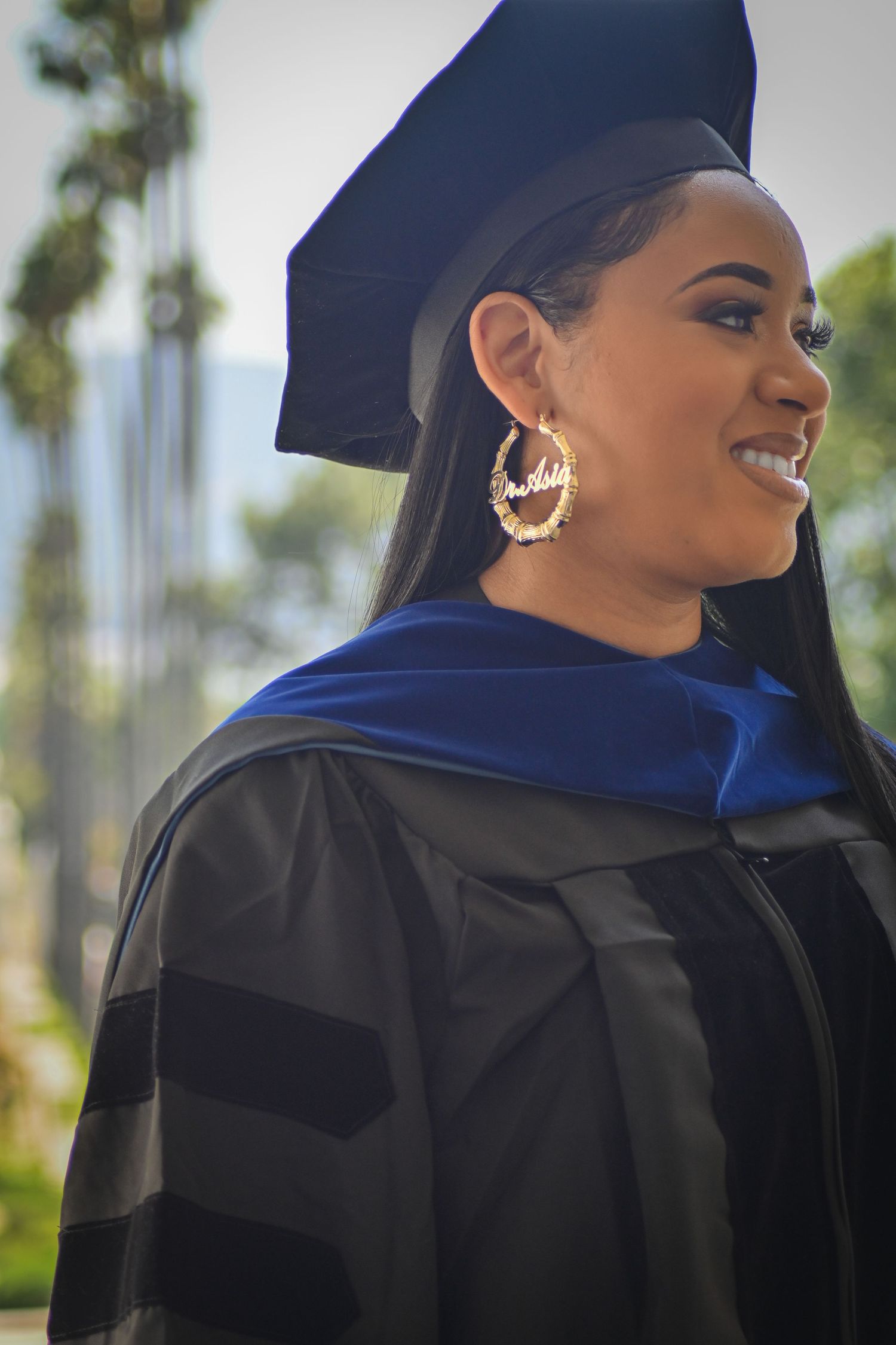 Graduate in traditional cap and gown smiling while wearing ornate gold hoop earrings.
