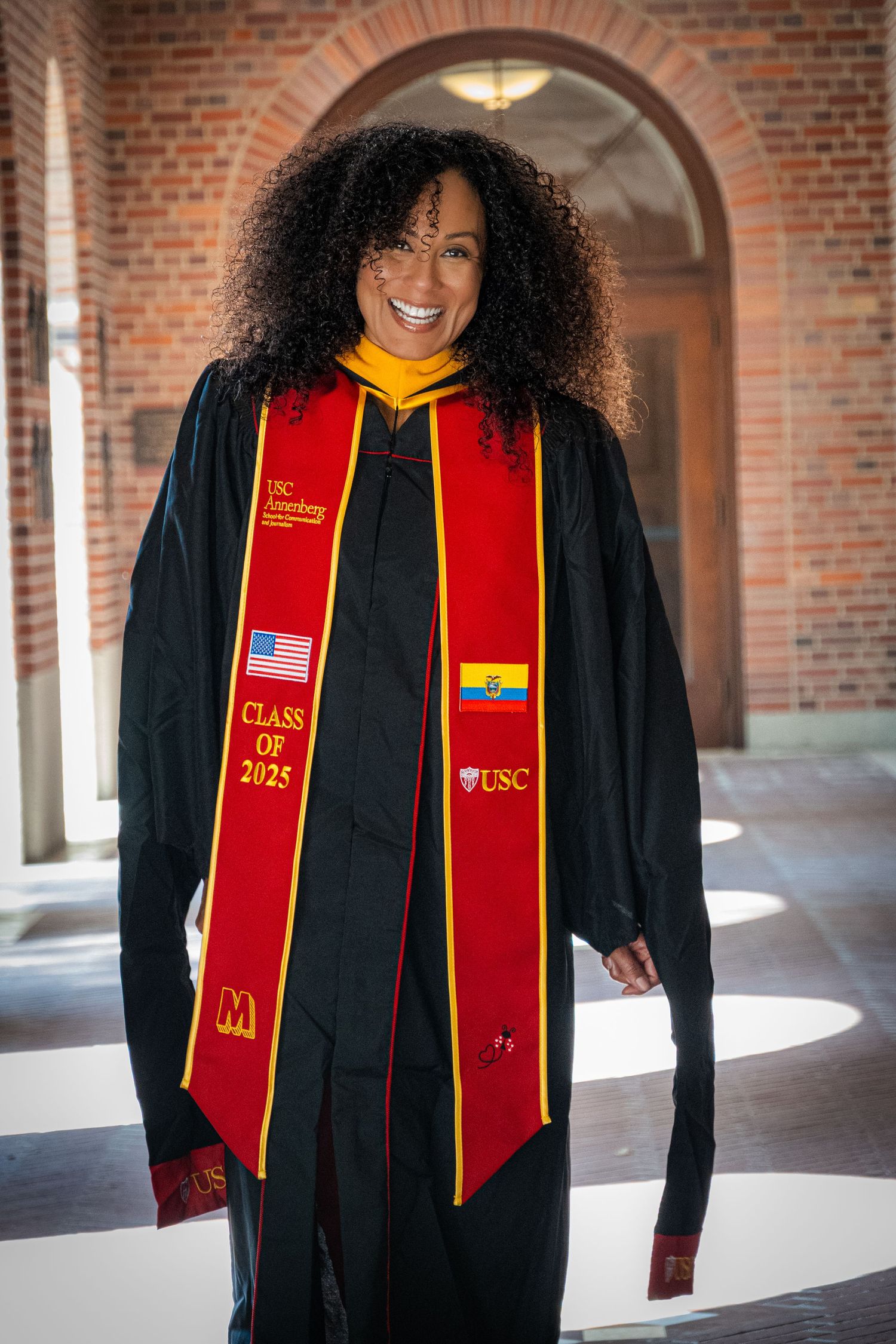 Graduate in USC Class of 2025 regalia smiles while standing in arched brick corridor.
