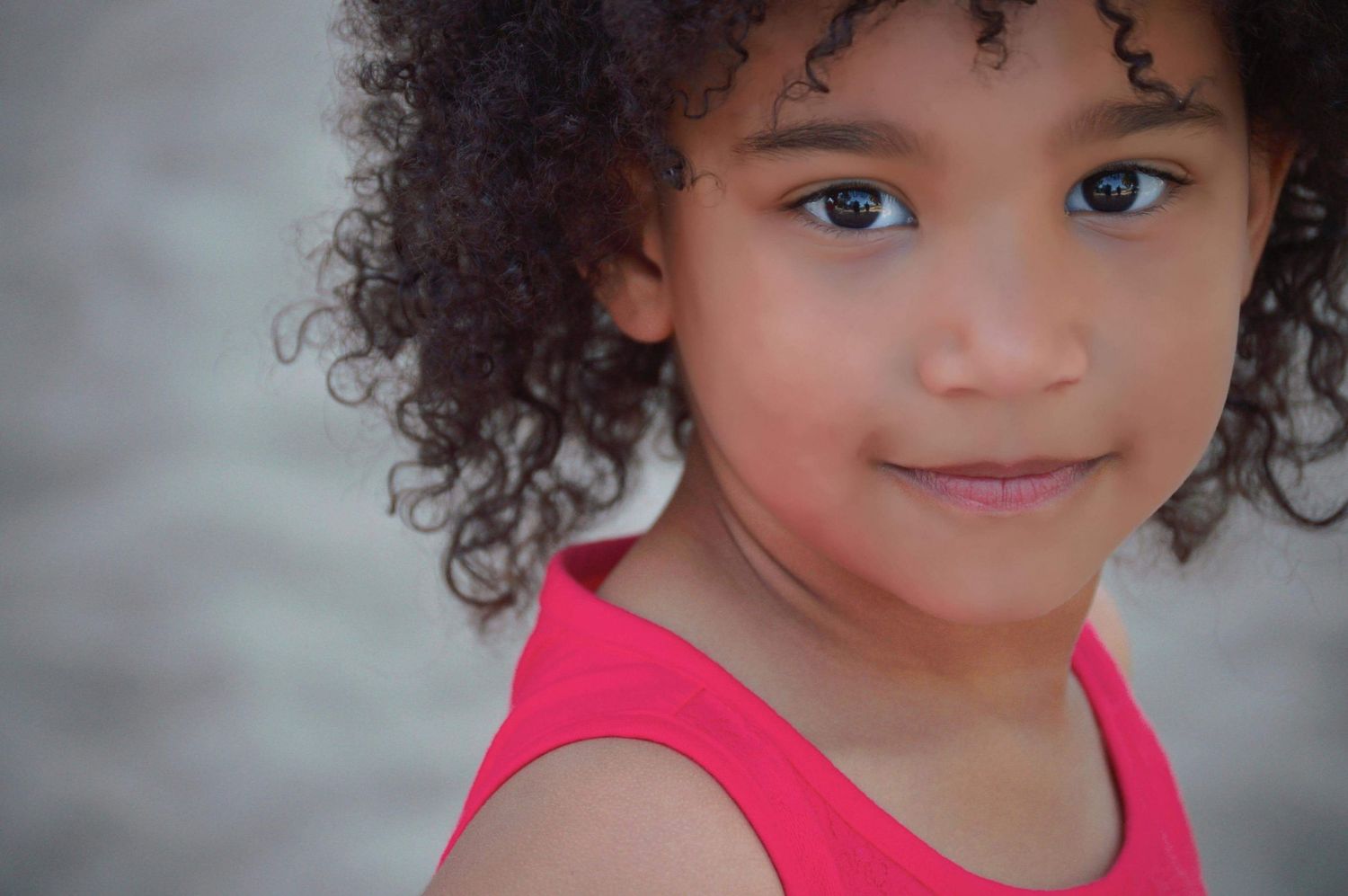 A young mixed-race girl with curly hair smiles softly, wearing a pink tank top against a light background.